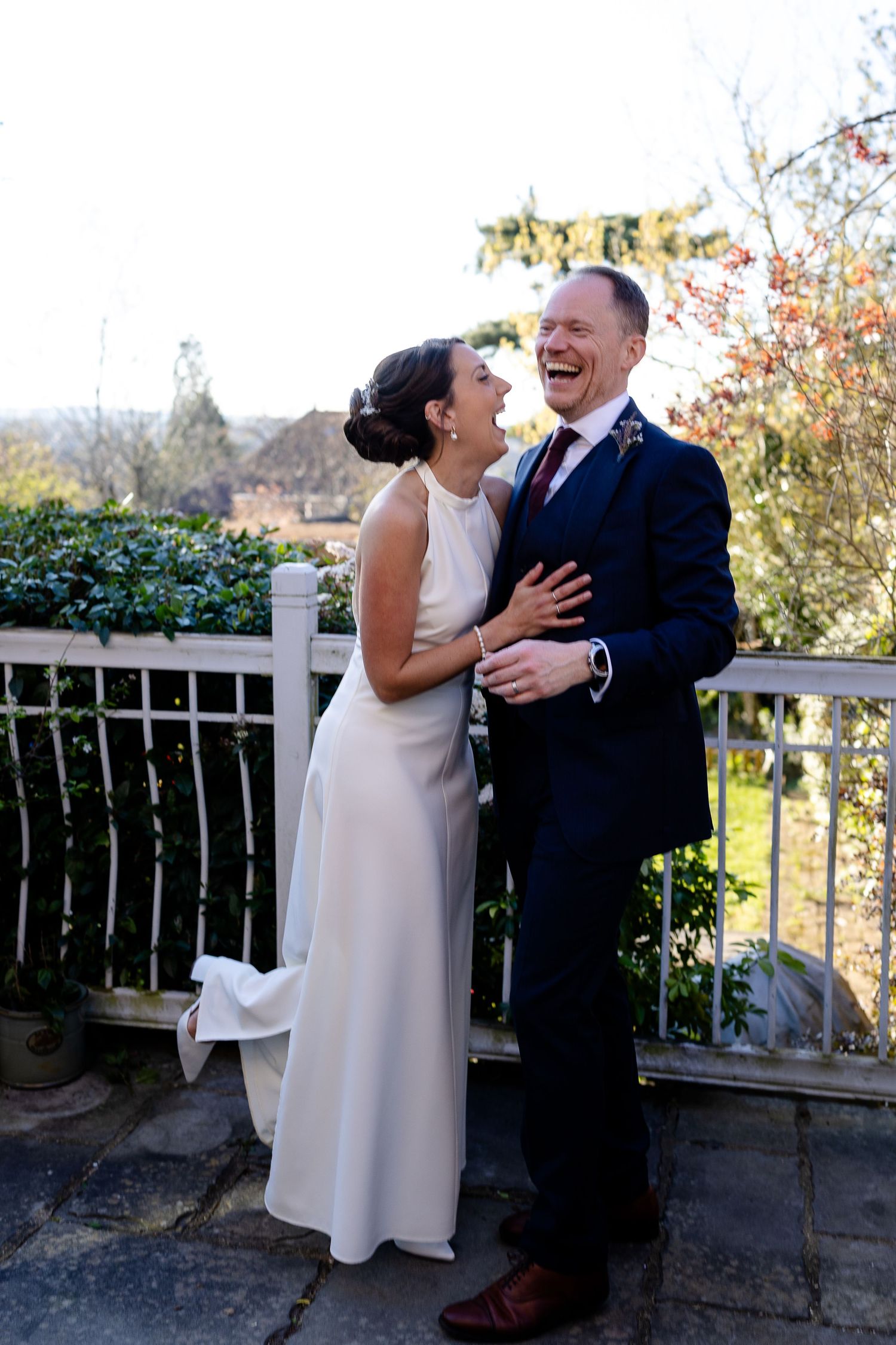 Bride and groom laughing joyfully together on a terrace, wearing wedding attire outdoors.