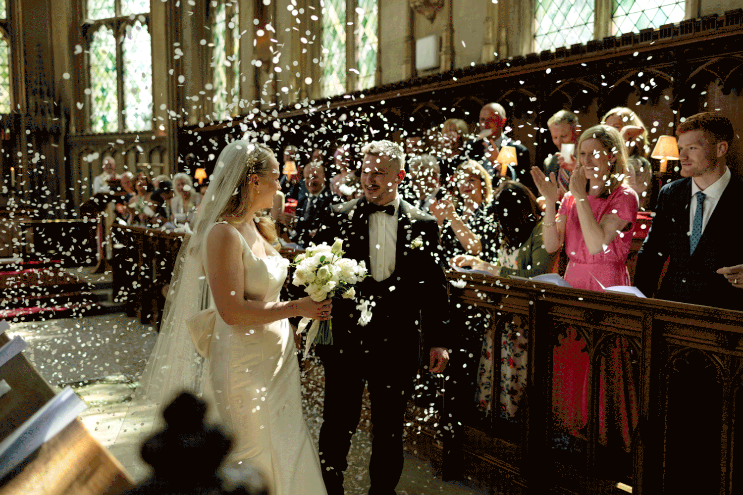 Confetti fills the air at an indoor wedding ceremony as a bride in white stands at the altar.
