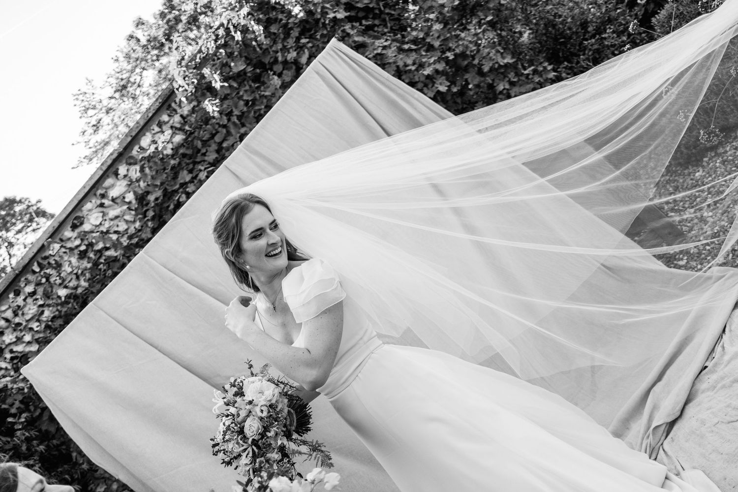 Black and white photo of a smiling bride in a short-sleeved gown holding a bouquet as her long veil billows in the wind outdoors.