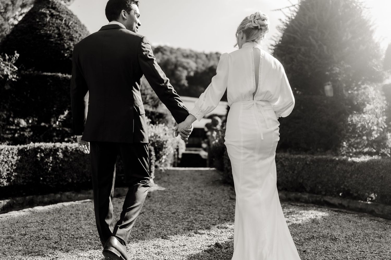 Black and white photo of a bride and groom walking hand in hand through a garden on their wedding day.