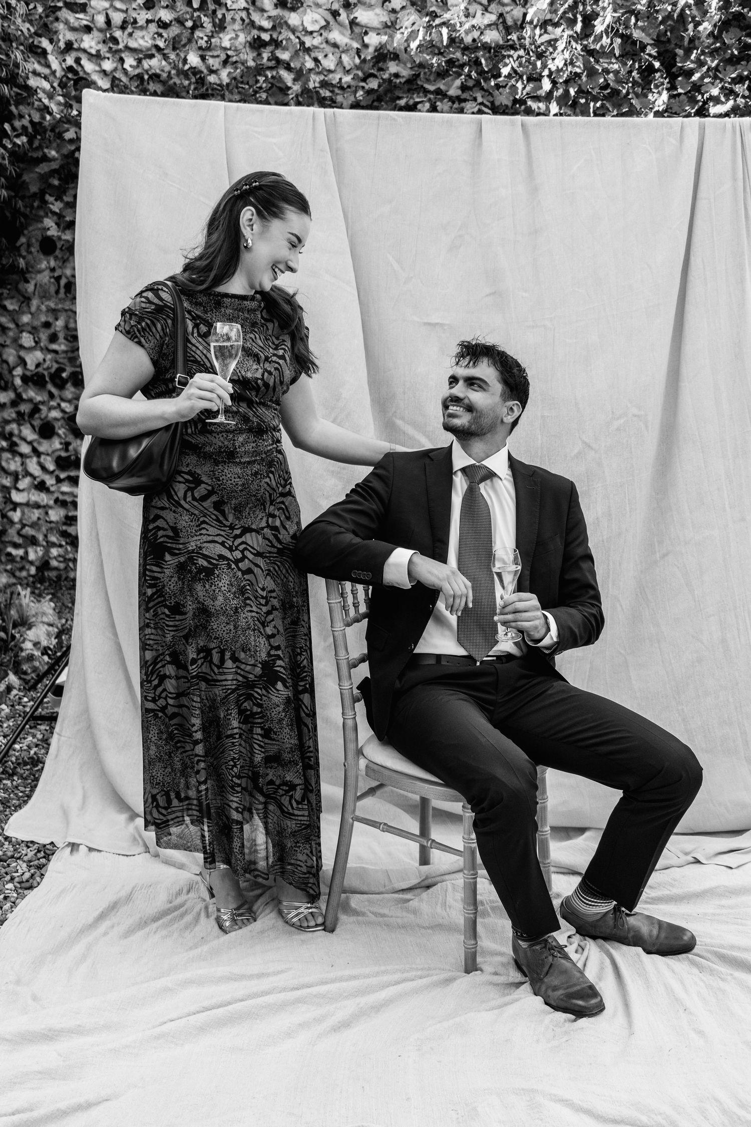 Black and white photo of a smiling woman in a lace dress standing beside a seated man in a suit, both holding champagne glasses.