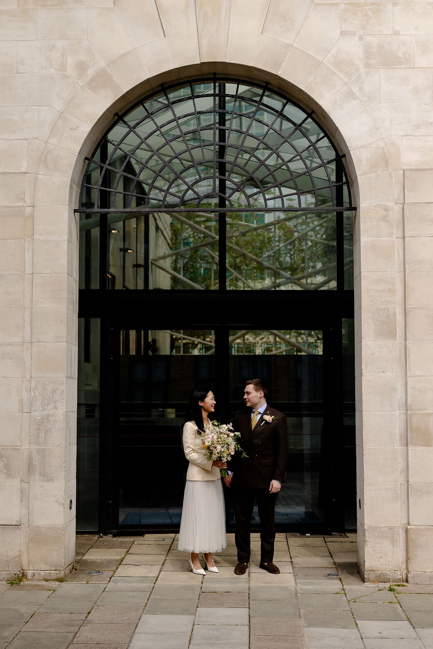 Bride and groom stand before grand arched glass door at elegant stone building wedding venue.