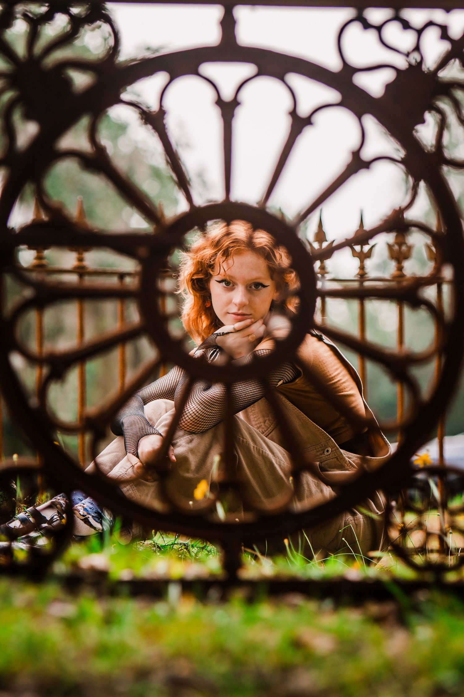 Creative portrait through decorative iron gate with floral pattern creating circular frame against natural outdoor background.
