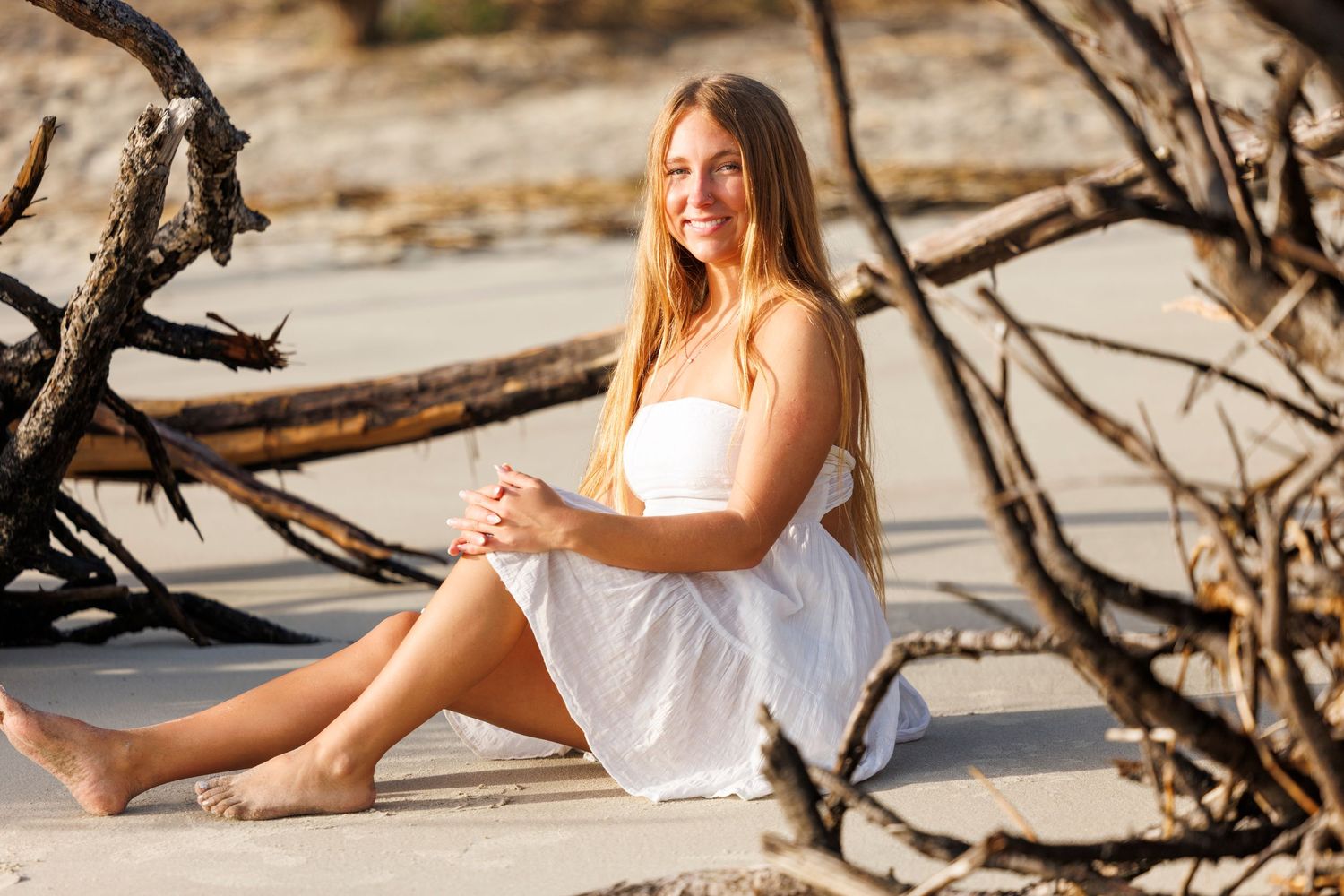 A woman in a white dress poses gracefully on a sandy beach with driftwood creating a natural backdrop.