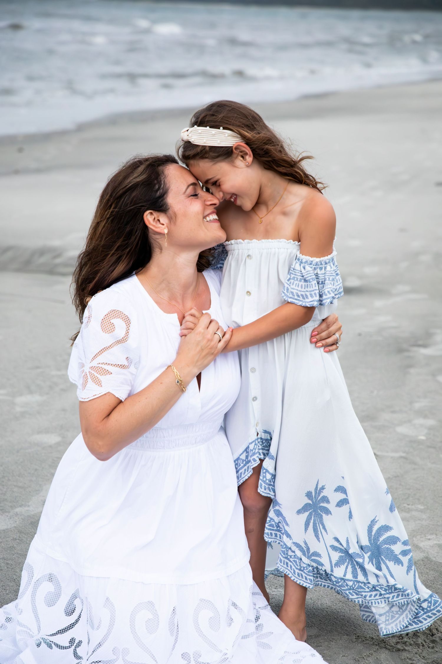 Two people in white dresses share an intimate moment on a beach, with waves visible in the background.