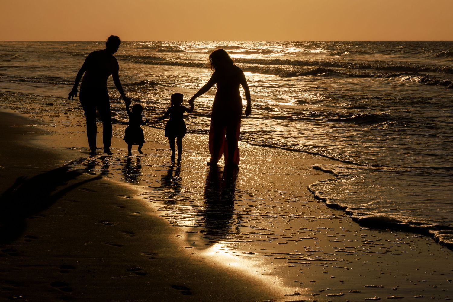Silhouetted figures walk along a golden beach at sunset with waves gently rolling onto the shore.