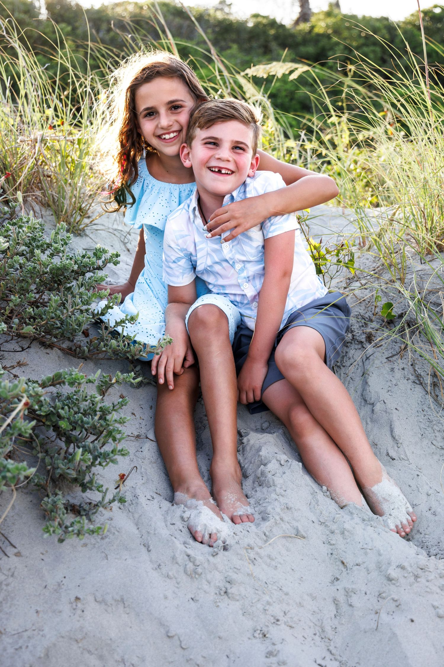 Two children in matching blue and white outfits sit together on a sandy beach dune during a summer photoshoot.