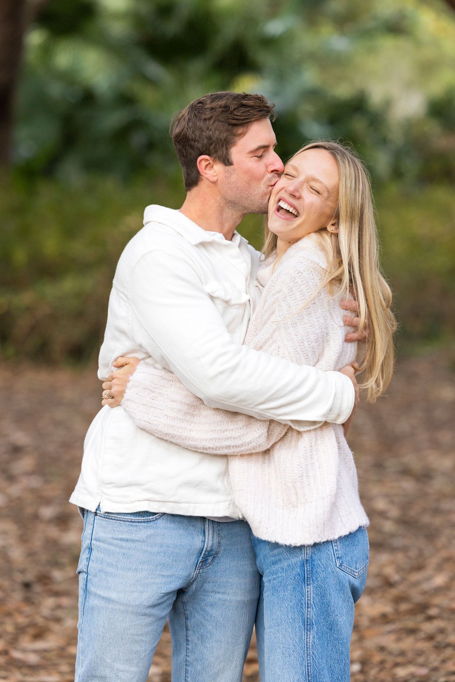 A couple wearing white sweaters and jeans share a joyful embrace and laugh together outdoors.