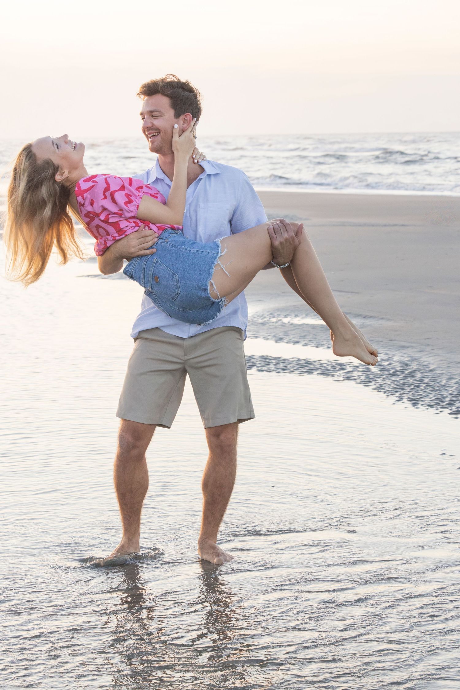 A couple plays in the shallow ocean waves at sunset while dipping their feet in the water on a beach.