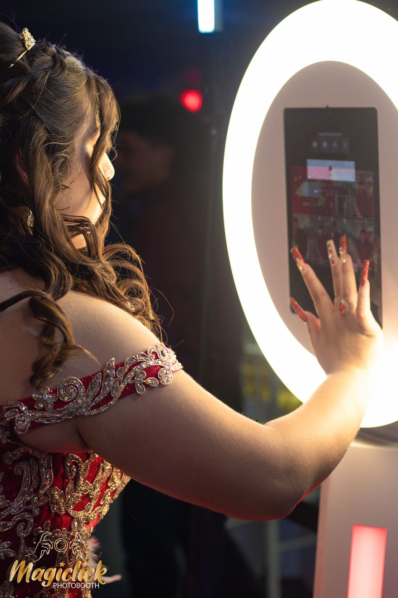 A person in an elegant red and white dress poses next to a large illuminated ring light in a dark setting.
