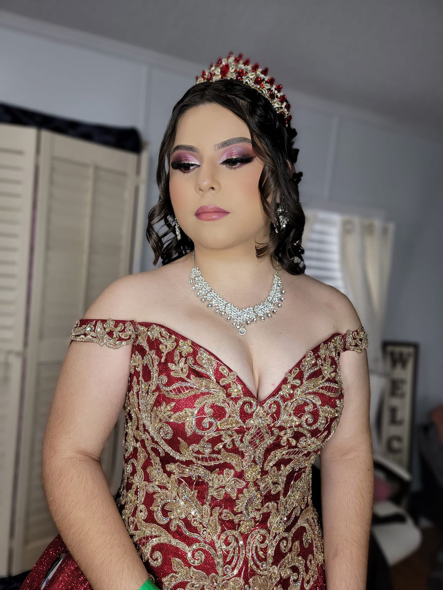 Elegant formal gown features intricate red and gold embroidery with off-shoulder design, paired with crystal crown and necklace.