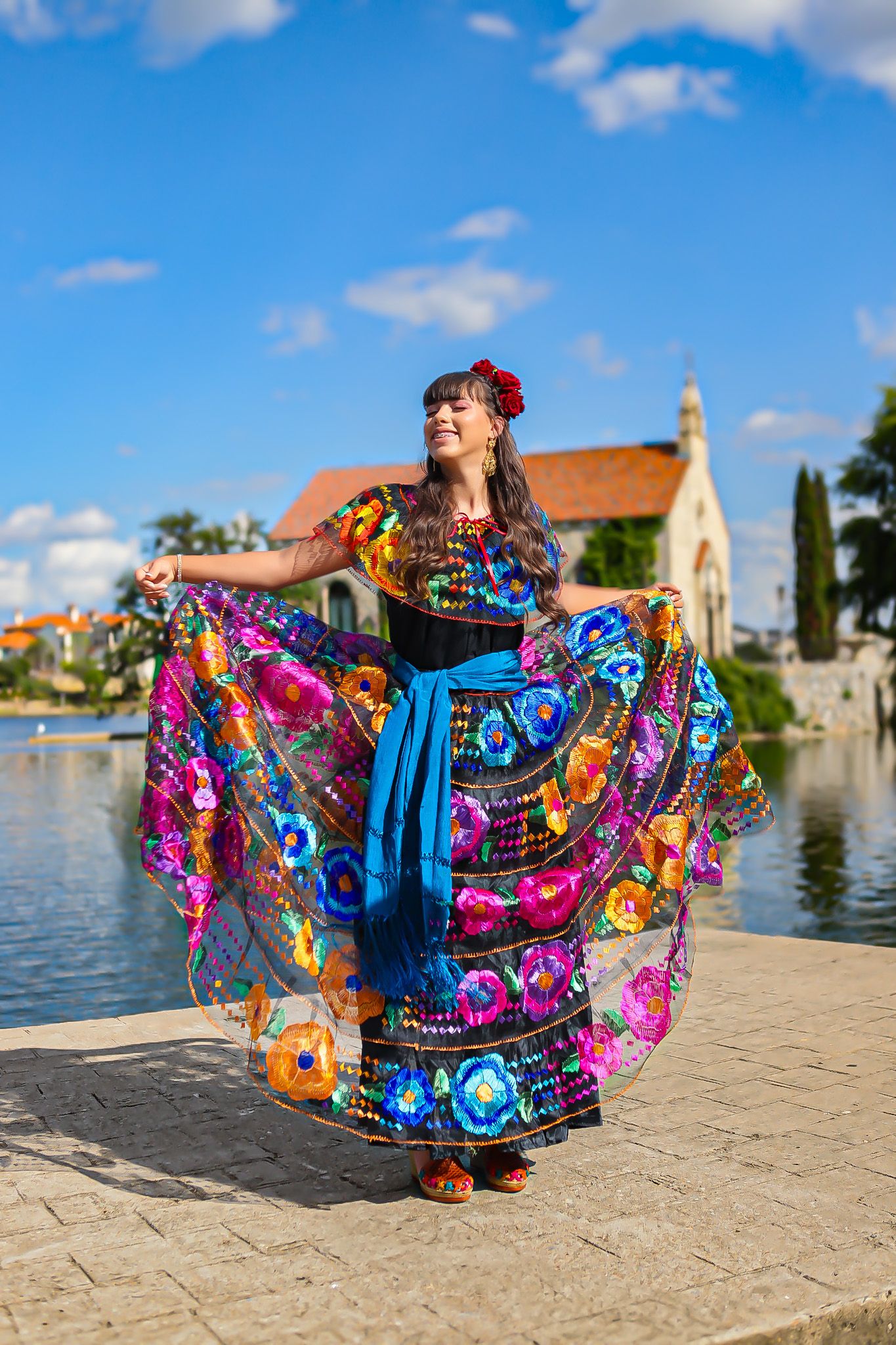 A colorful traditional Mexican dress with vibrant patterns swirls by a lakeside church under blue skies.