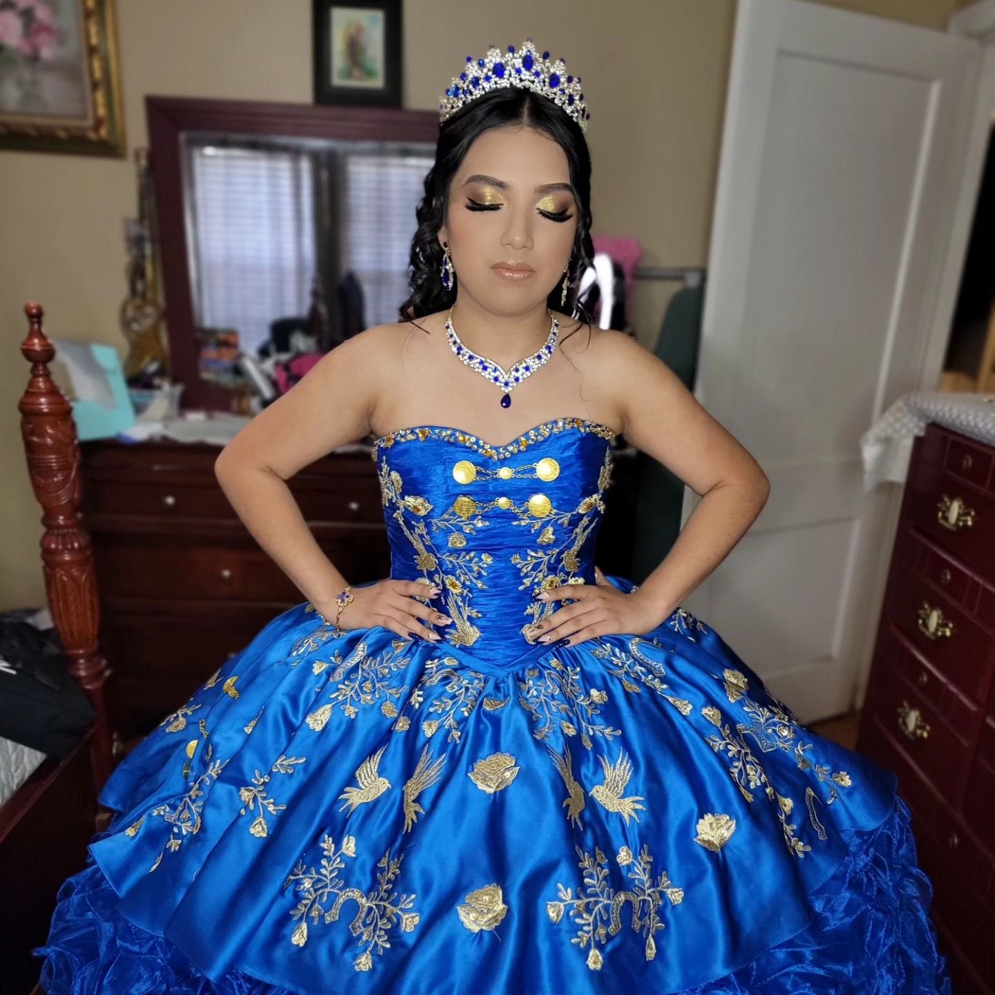 A person in a vibrant royal blue quinceañera ball gown with floral embellishments poses wearing a tiara and jewelry.