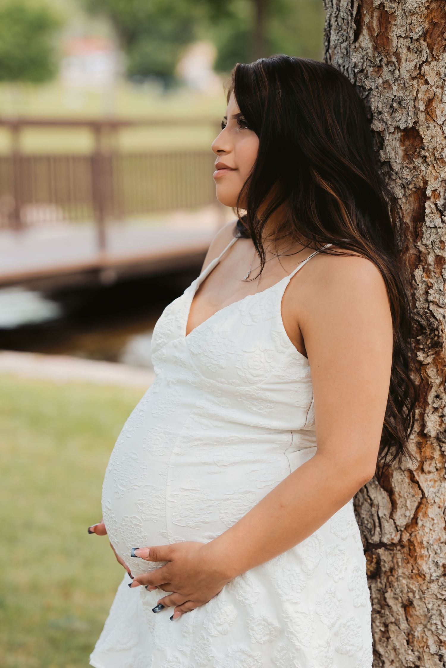 Pregnant woman in white lace dress leaning against tree in park, cradling baby bump, long dark hair, serene expression.