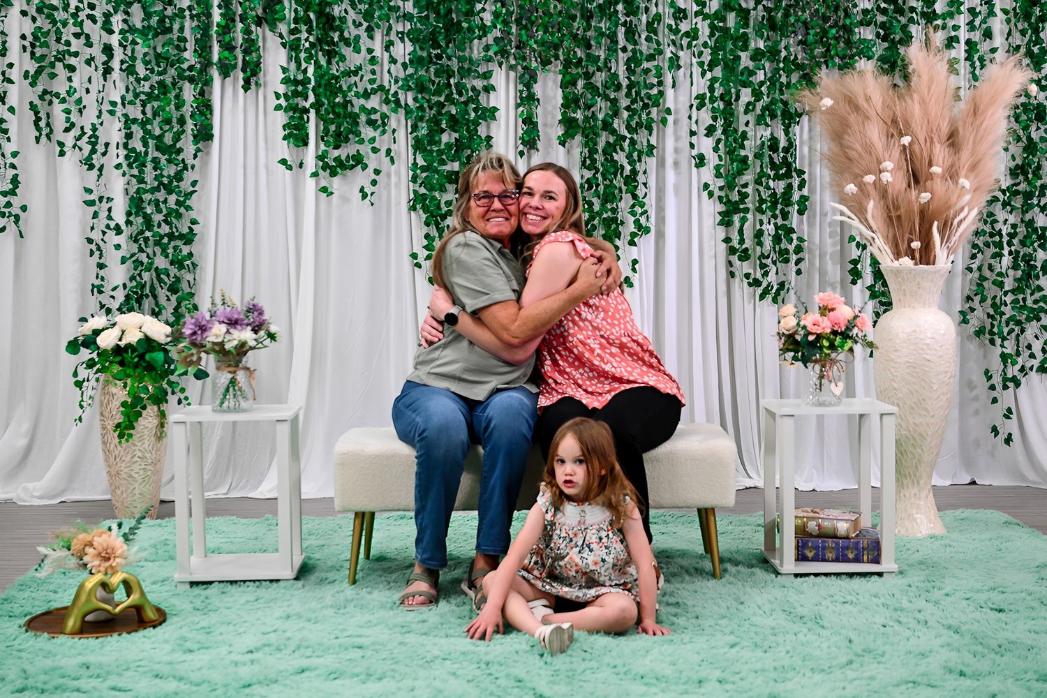 Two women hugging and a young girl sitting on green carpet before a floral backdrop with hanging greenery.