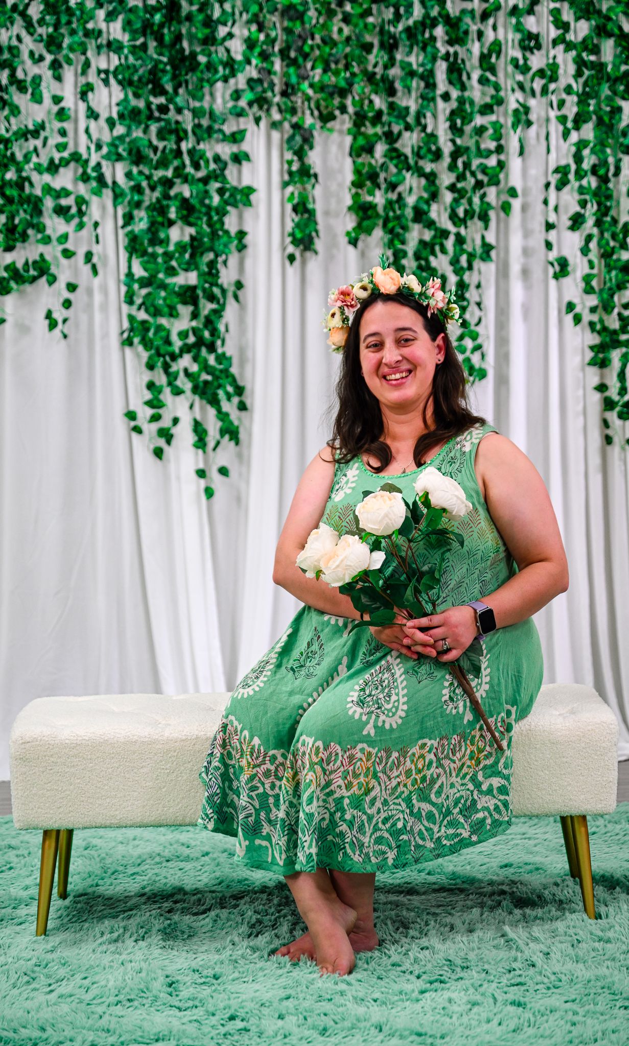 Woman in green floral dress holding white roses, wearing flower crown, seated on white bench with ivy backdrop.