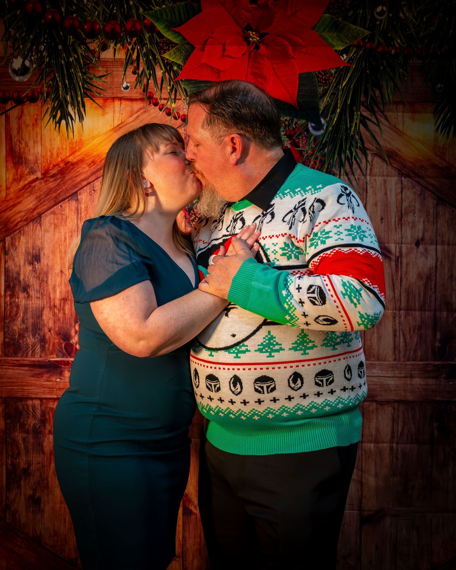 Couple sharing a kiss in front of Christmas decorations, man wearing festive ugly sweater, woman in teal dress.
