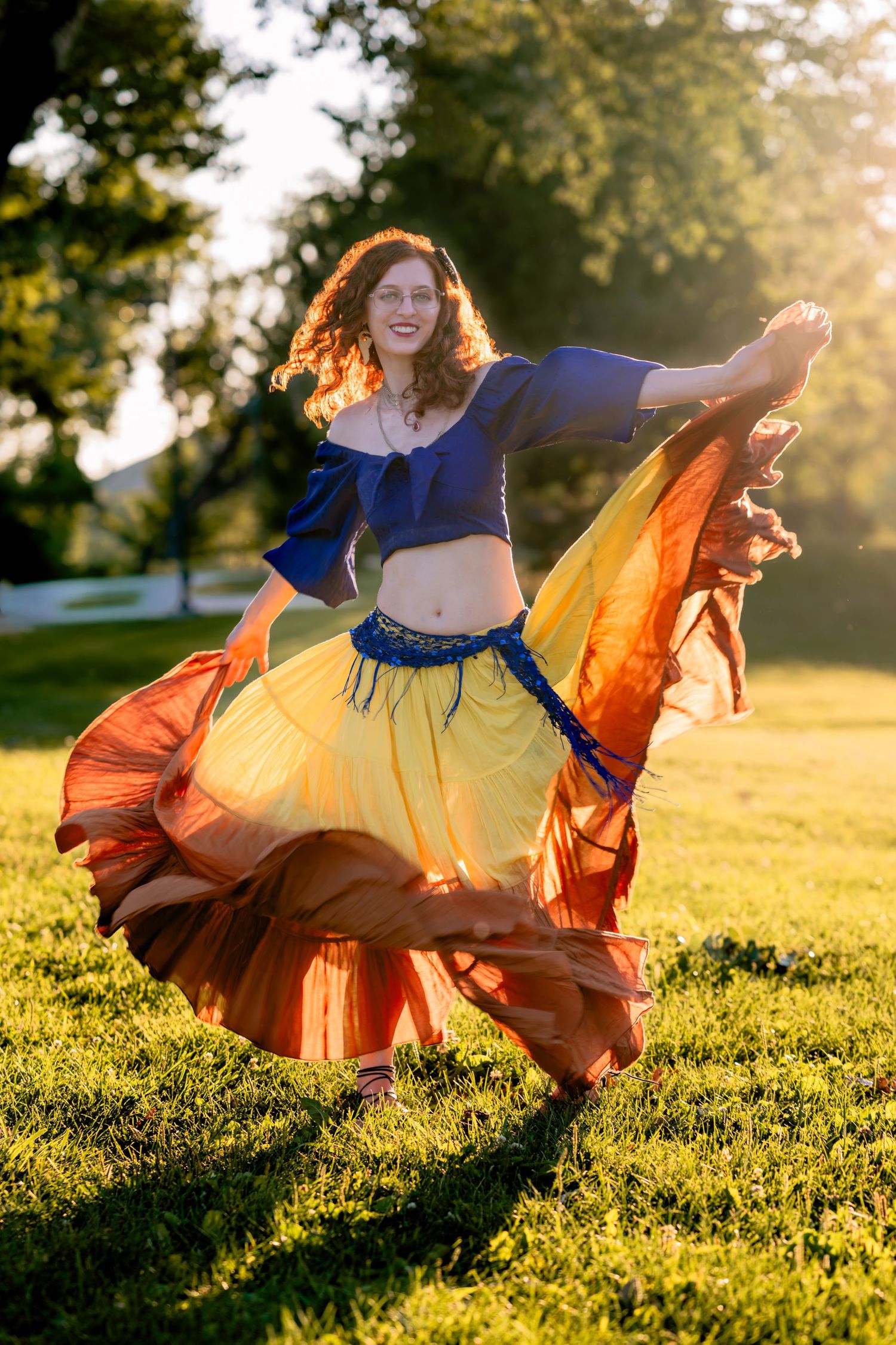 A redhead woman dances outdoors in a colorful yellow and orange skirt with a blue crop top in golden sunlight.