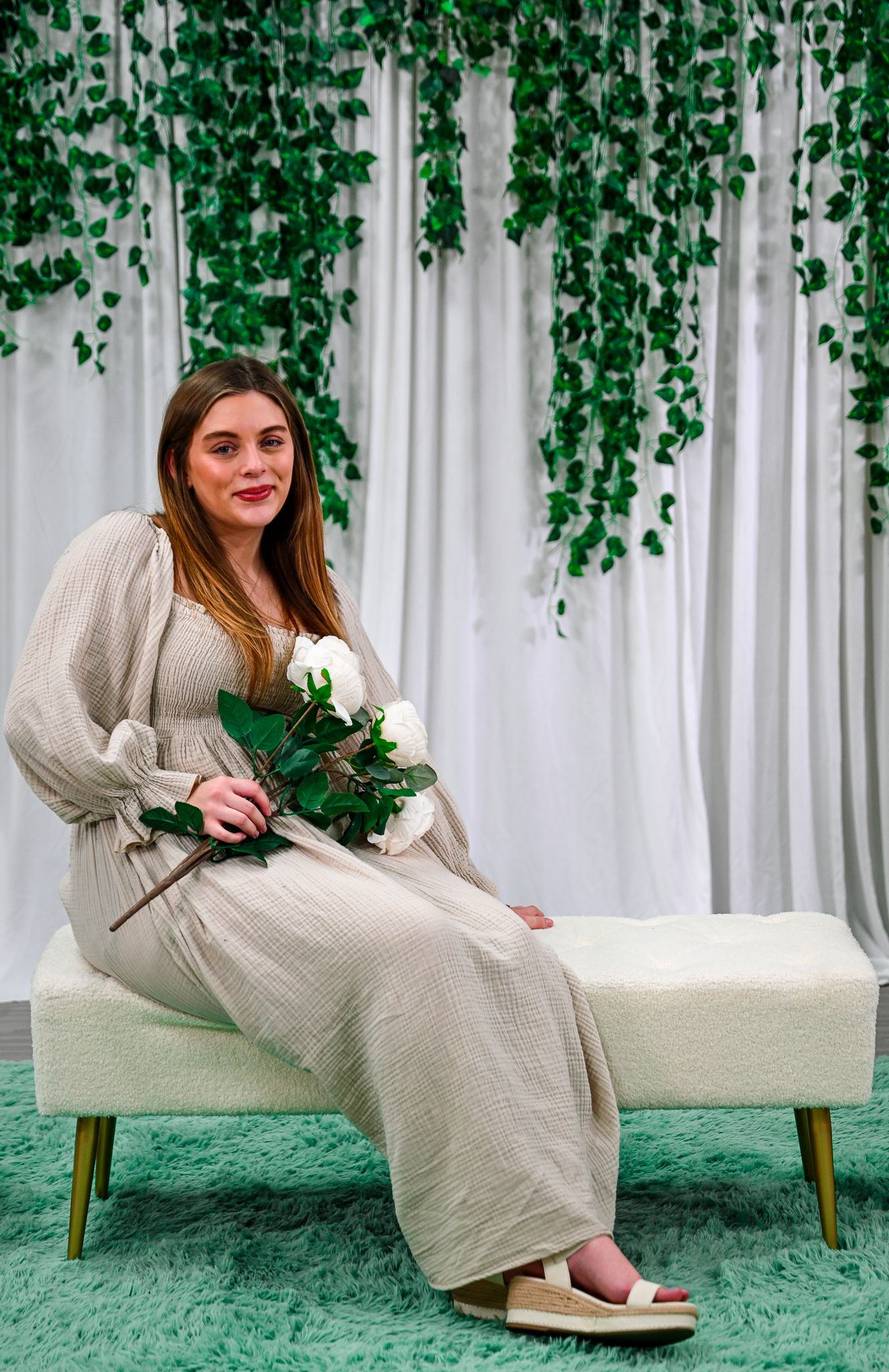 Woman in beige maxi dress holding white roses, seated on cream bench with green ivy backdrop.