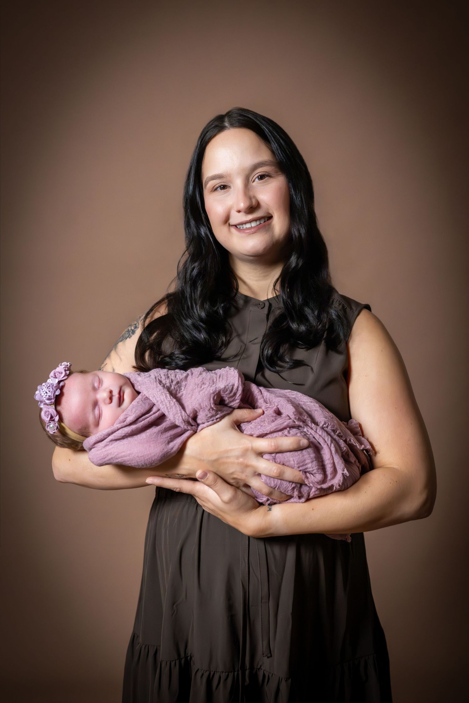 Smiling woman in dark dress holding a swaddled newborn baby in pink wrap and floral headband against brown backdrop.