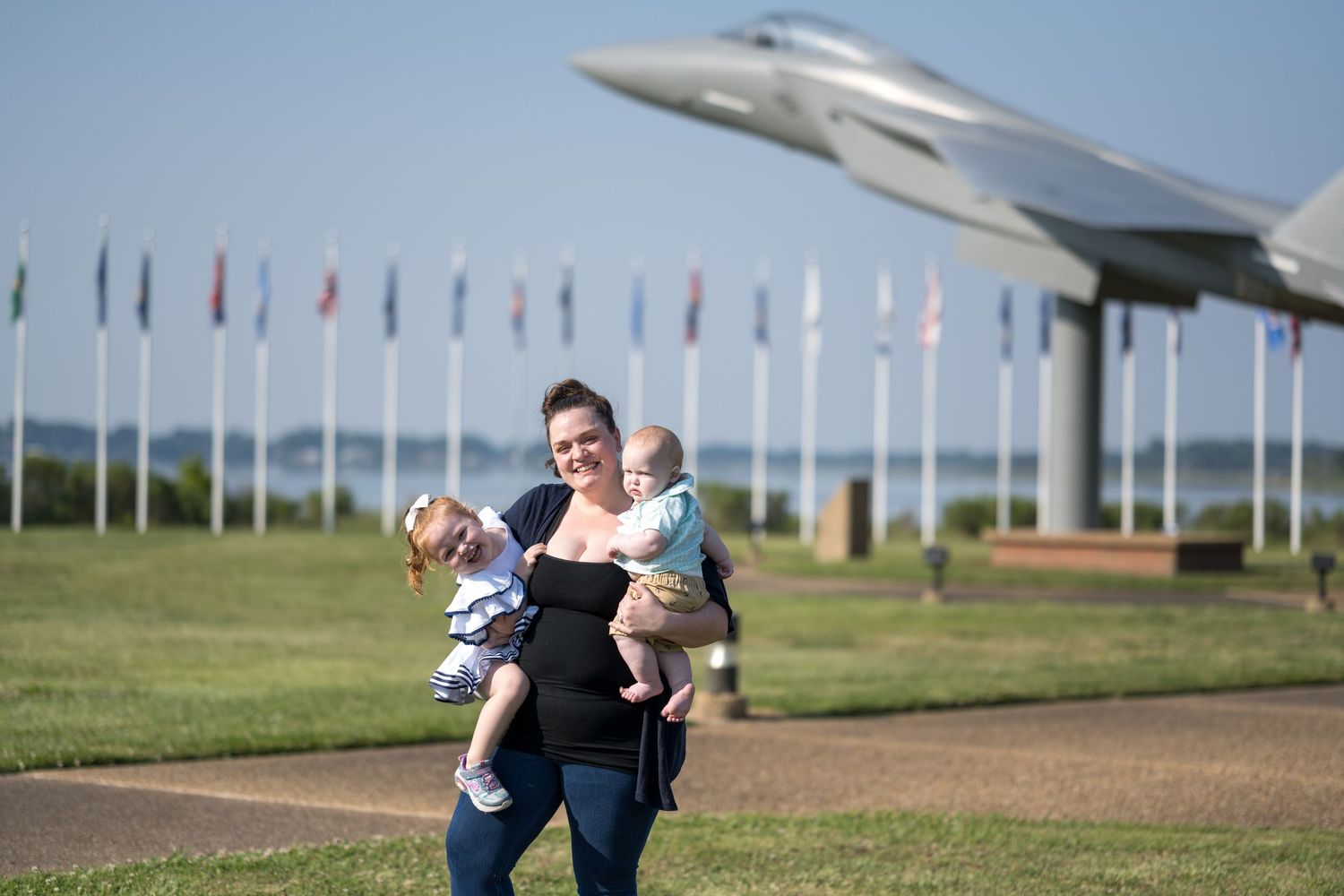 Woman carrying two young children near a military jet display with international flags at an air base.