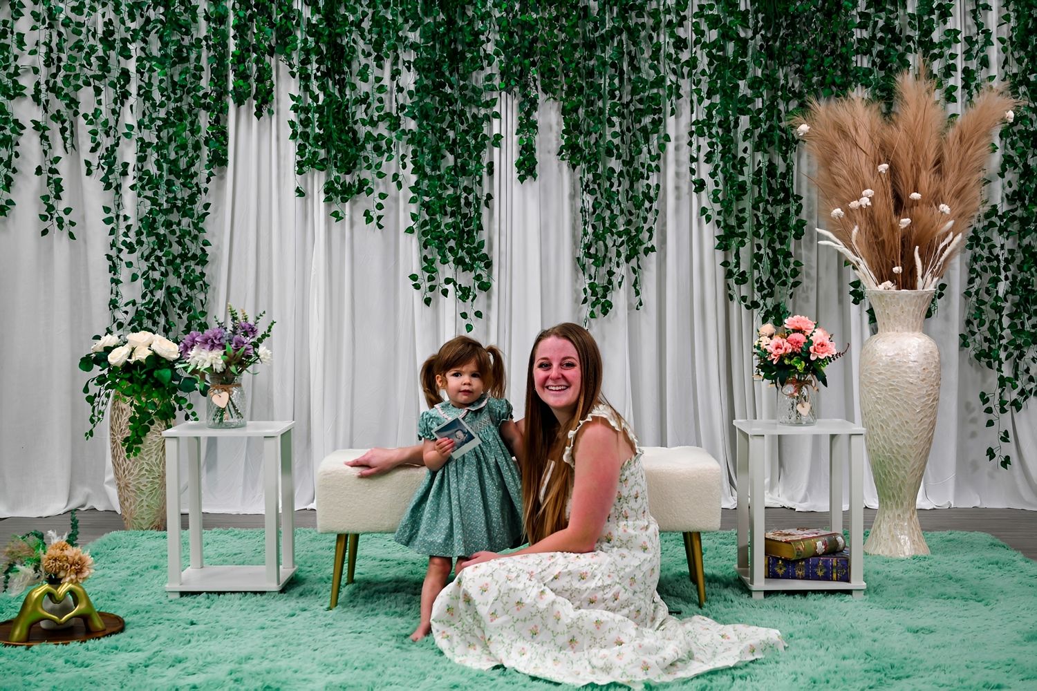 Woman and toddler girl in matching green dresses pose before elegant white backdrop with hanging greenery.