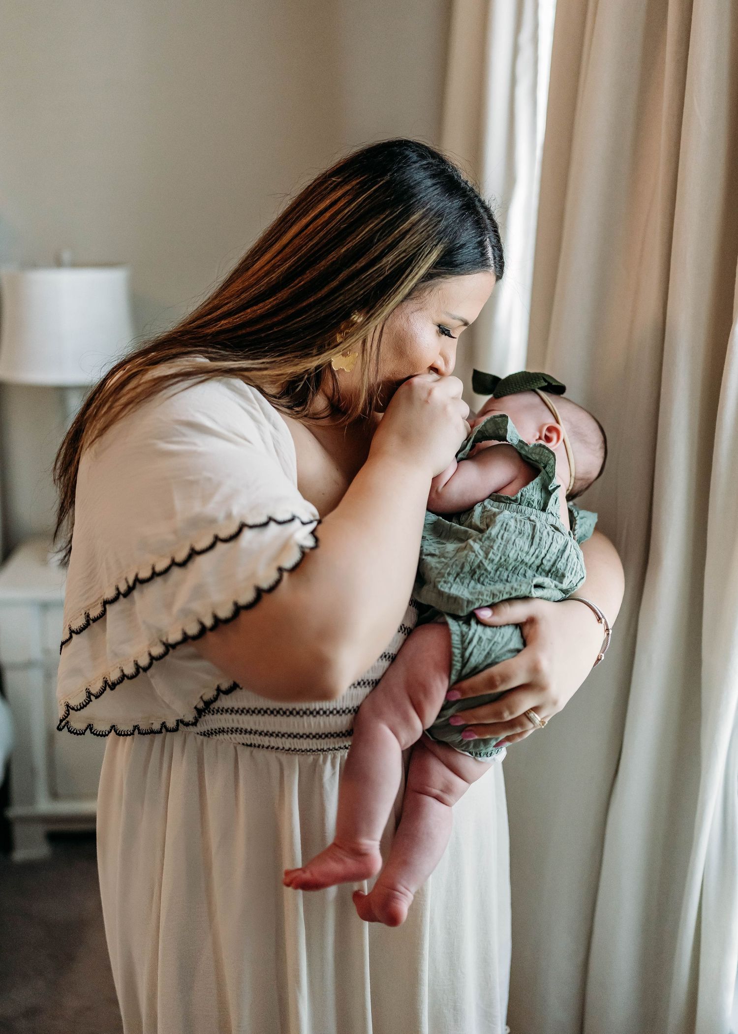 A tender moment by a window as a parent in a cream-colored dress holds and kisses their baby wearing sage green.