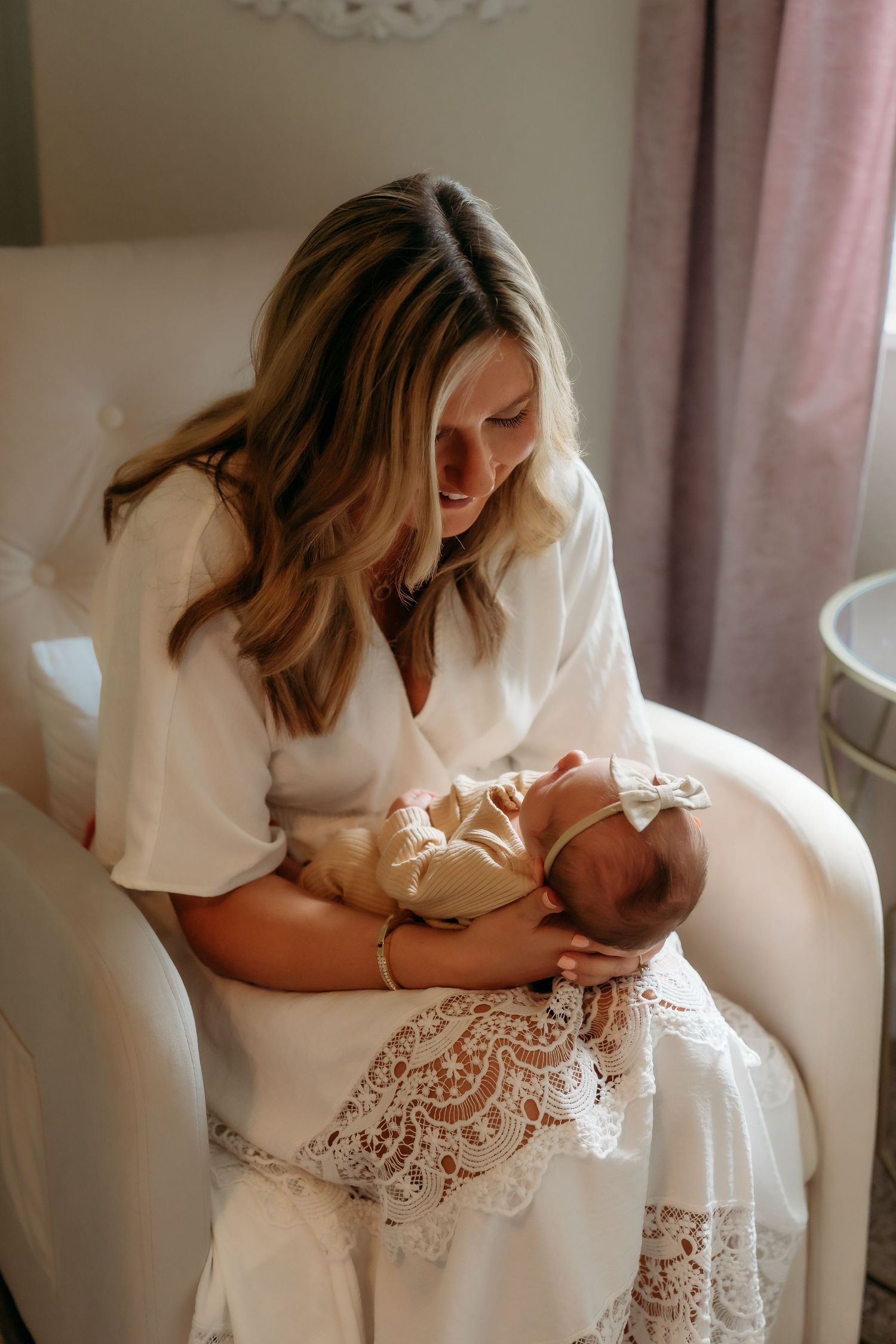 A woman in a white lace dress sits in a plush chair holding a newborn baby in a tender moment.