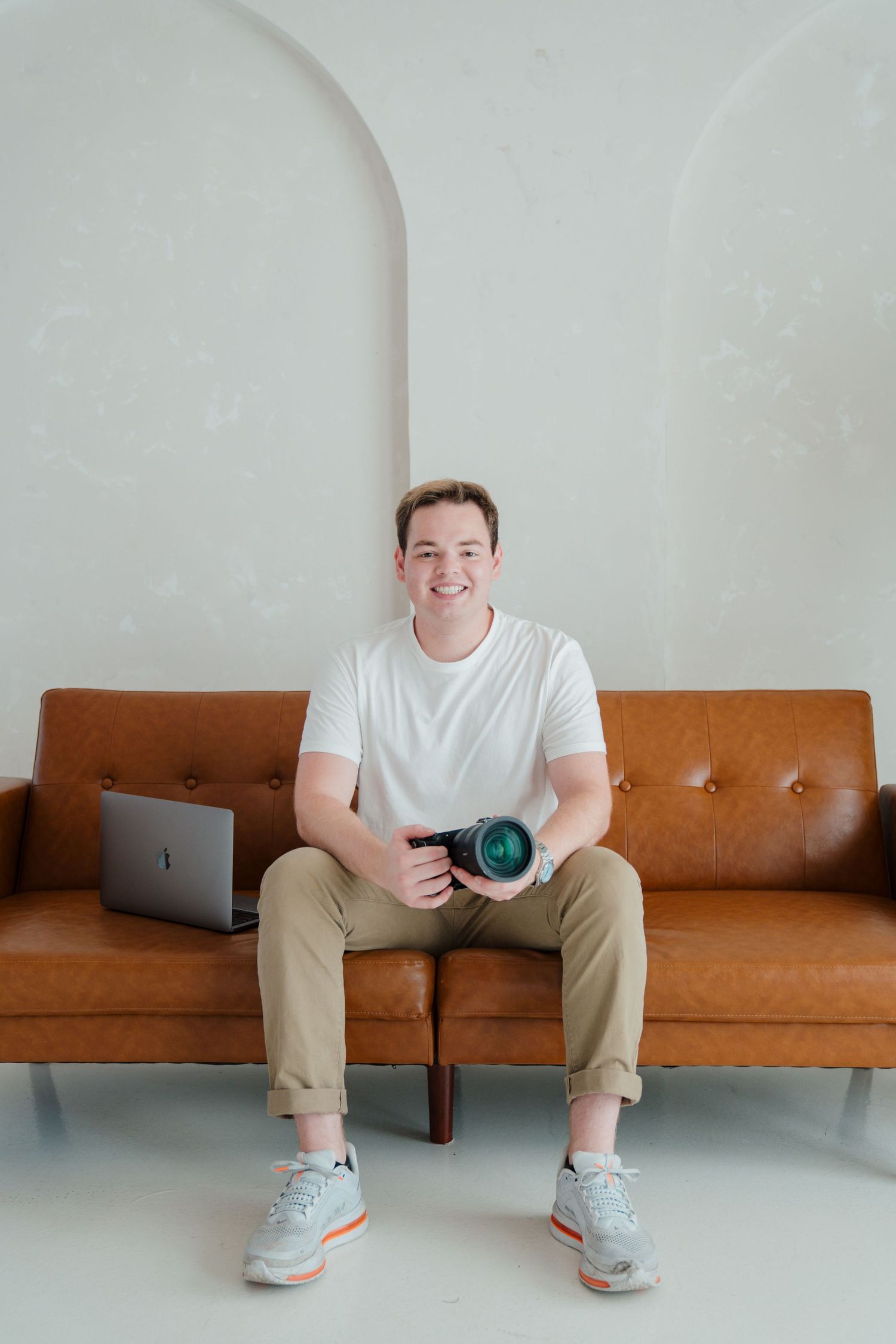 A photographer sits on a brown leather couch with a camera lens and laptop in a minimalist white room.