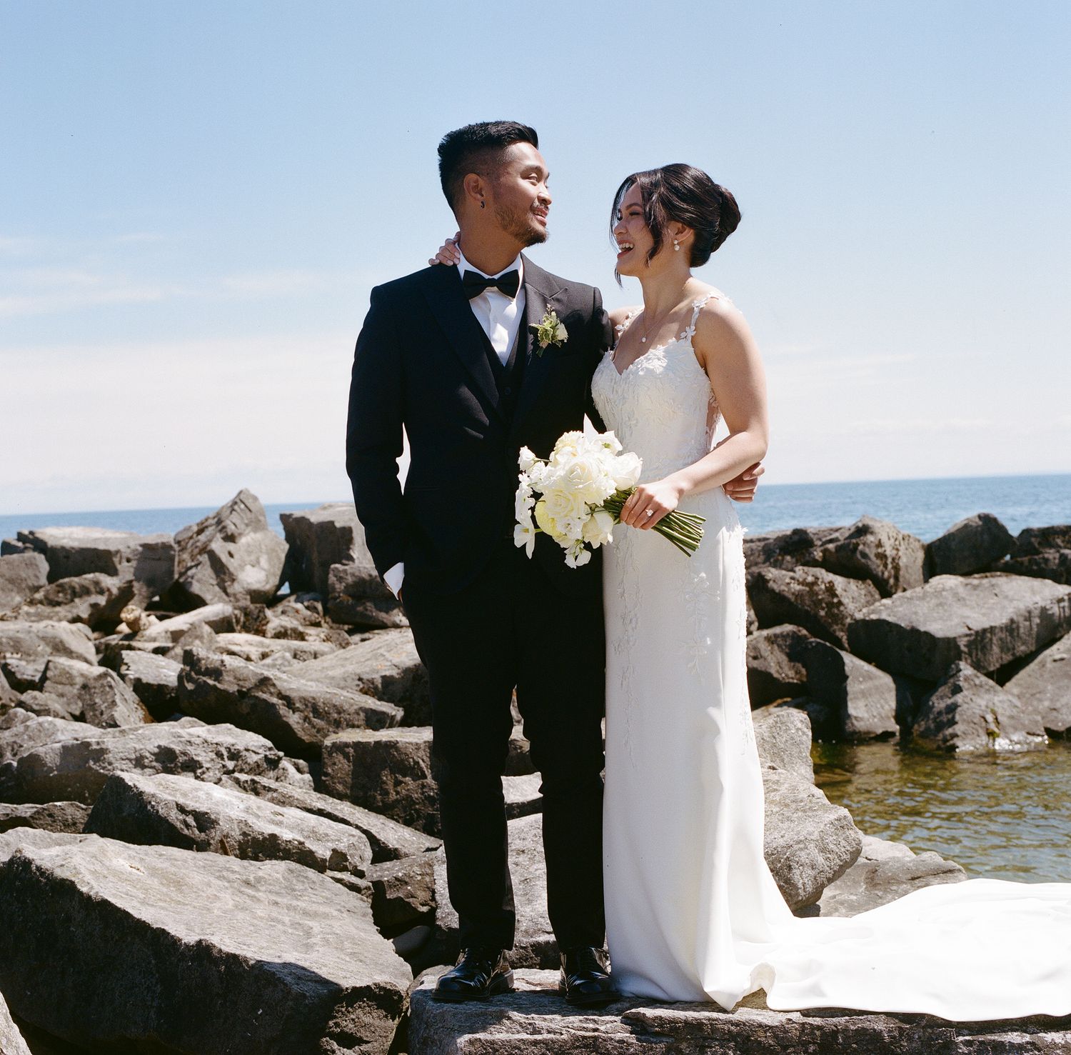 A newlywed couple shares a joyful moment together on rocky coastal terrain while holding a white floral bouquet.