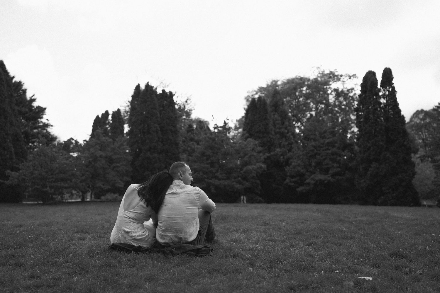 Black and white photo of couple sitting close together on grass in a tree-lined park.