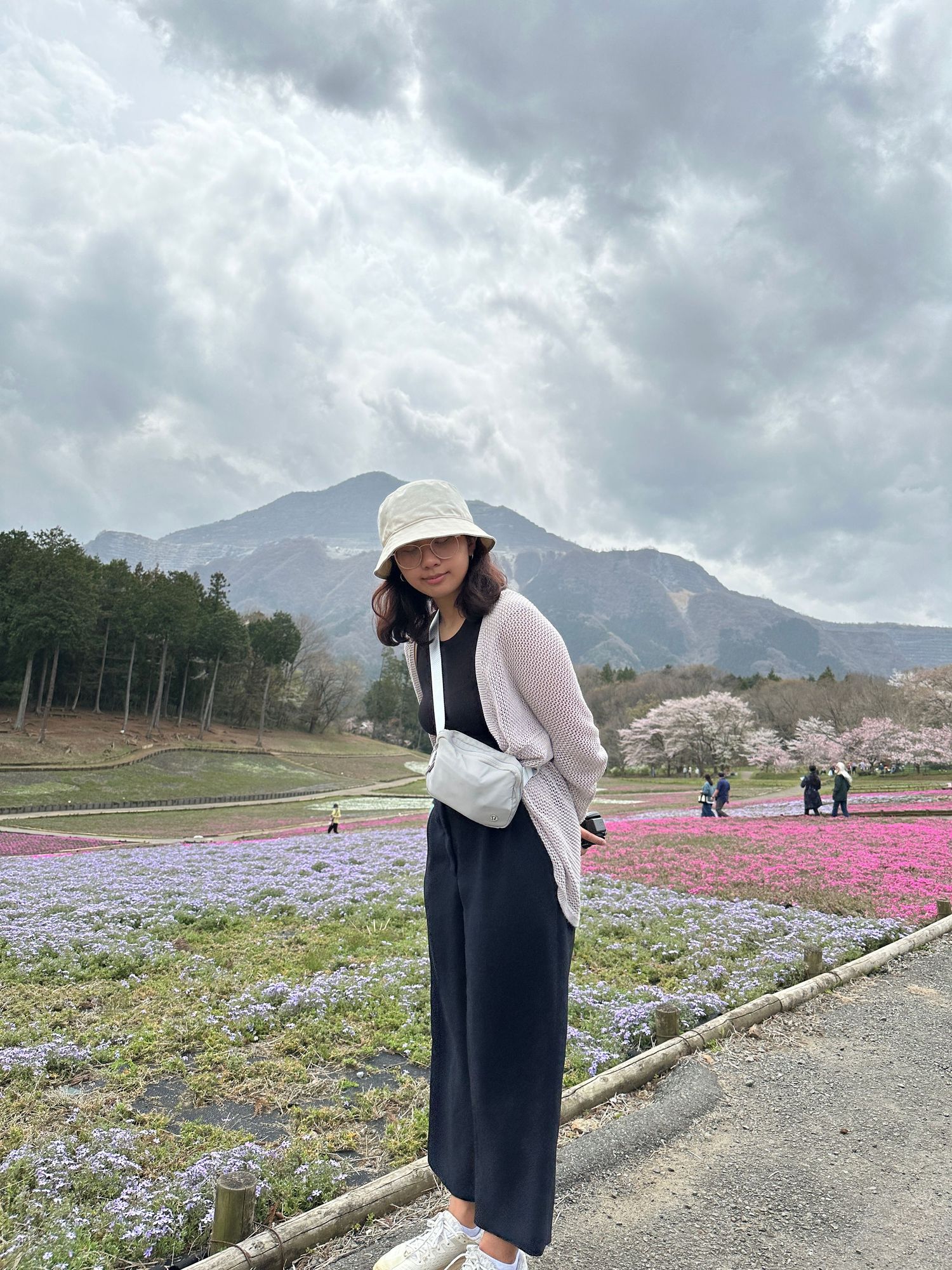 Woman in bucket hat and navy jumpsuit poses among pink moss phlox fields with cherry blossoms and mountain backdrop.
