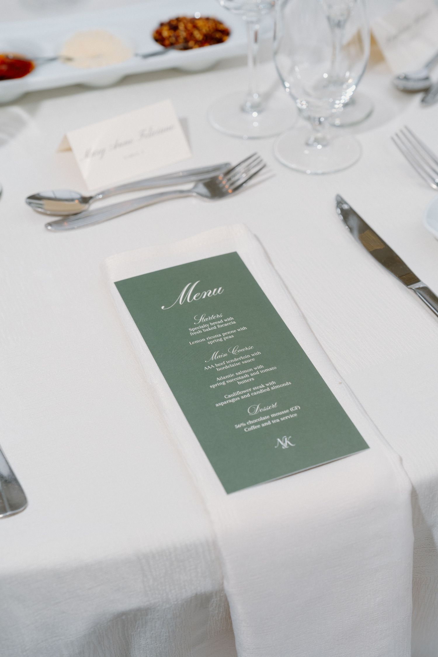 Elegant wedding table setting with green menu card on white linen, flanked by silverware and crystal glasses.