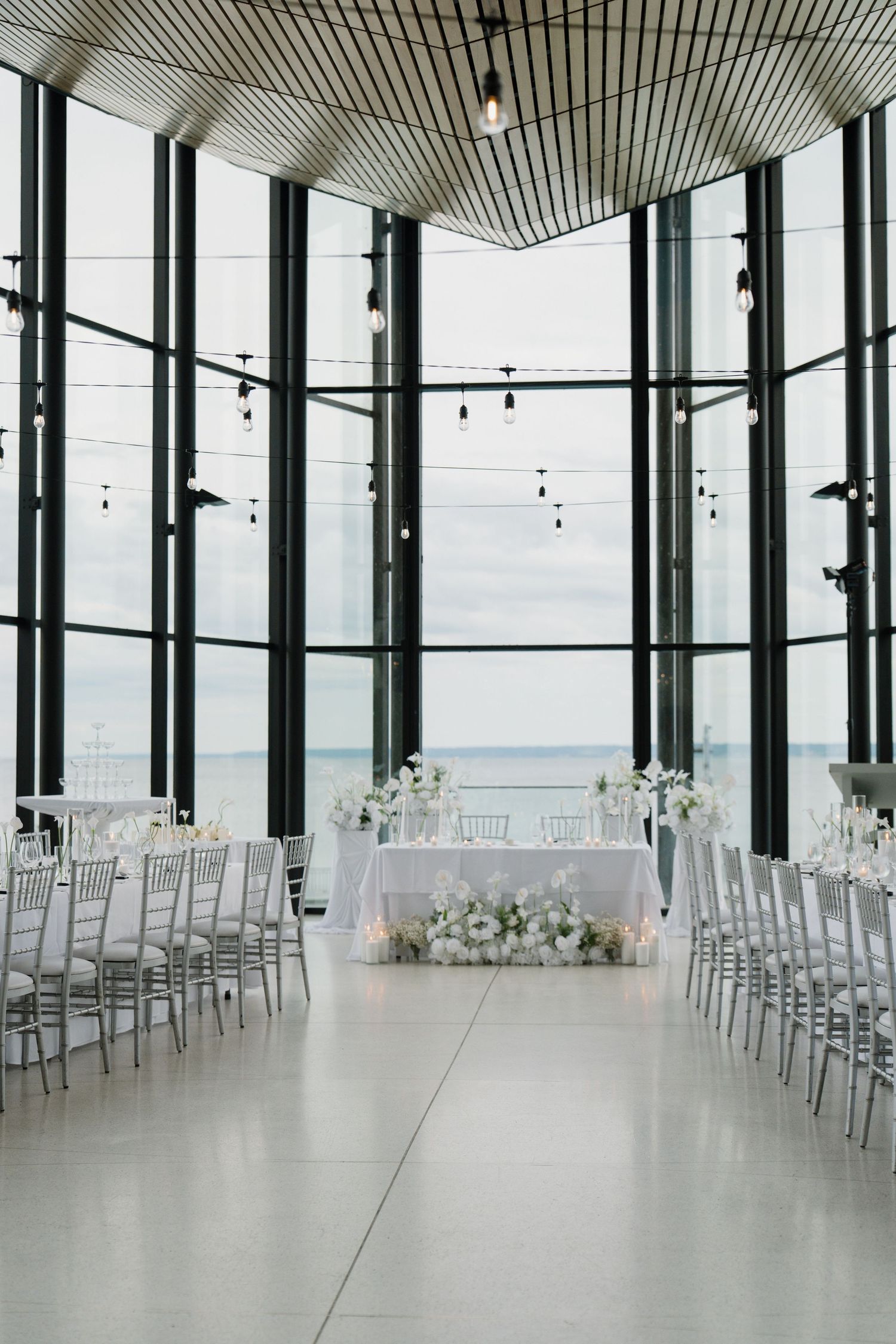 Elegant wedding ceremony setup with white chairs and floral arrangements in a modern venue with floor-to-ceiling ocean views.