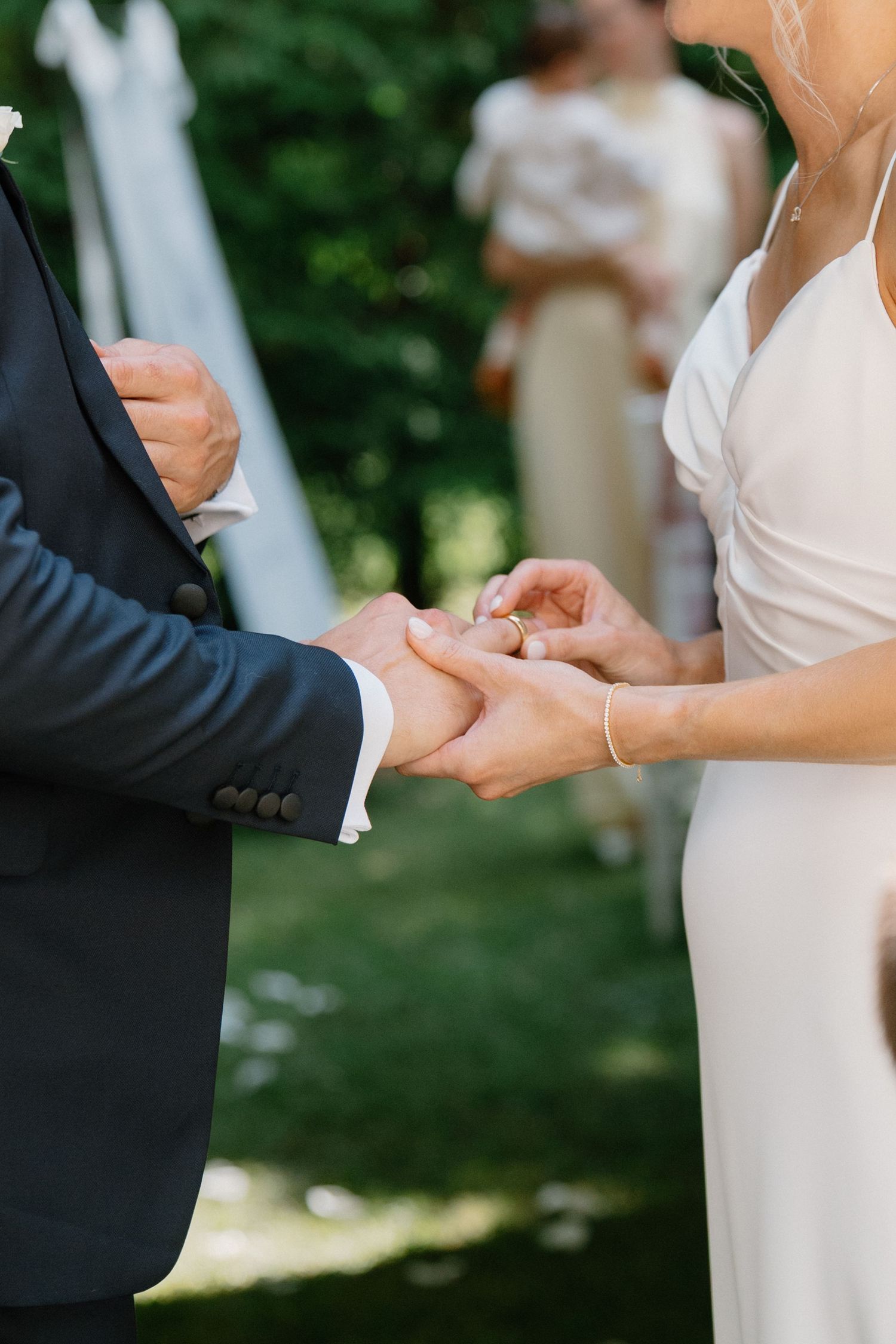 Bride placing a wedding ring on groom's finger during an outdoor ceremony, with guests visible in the background.