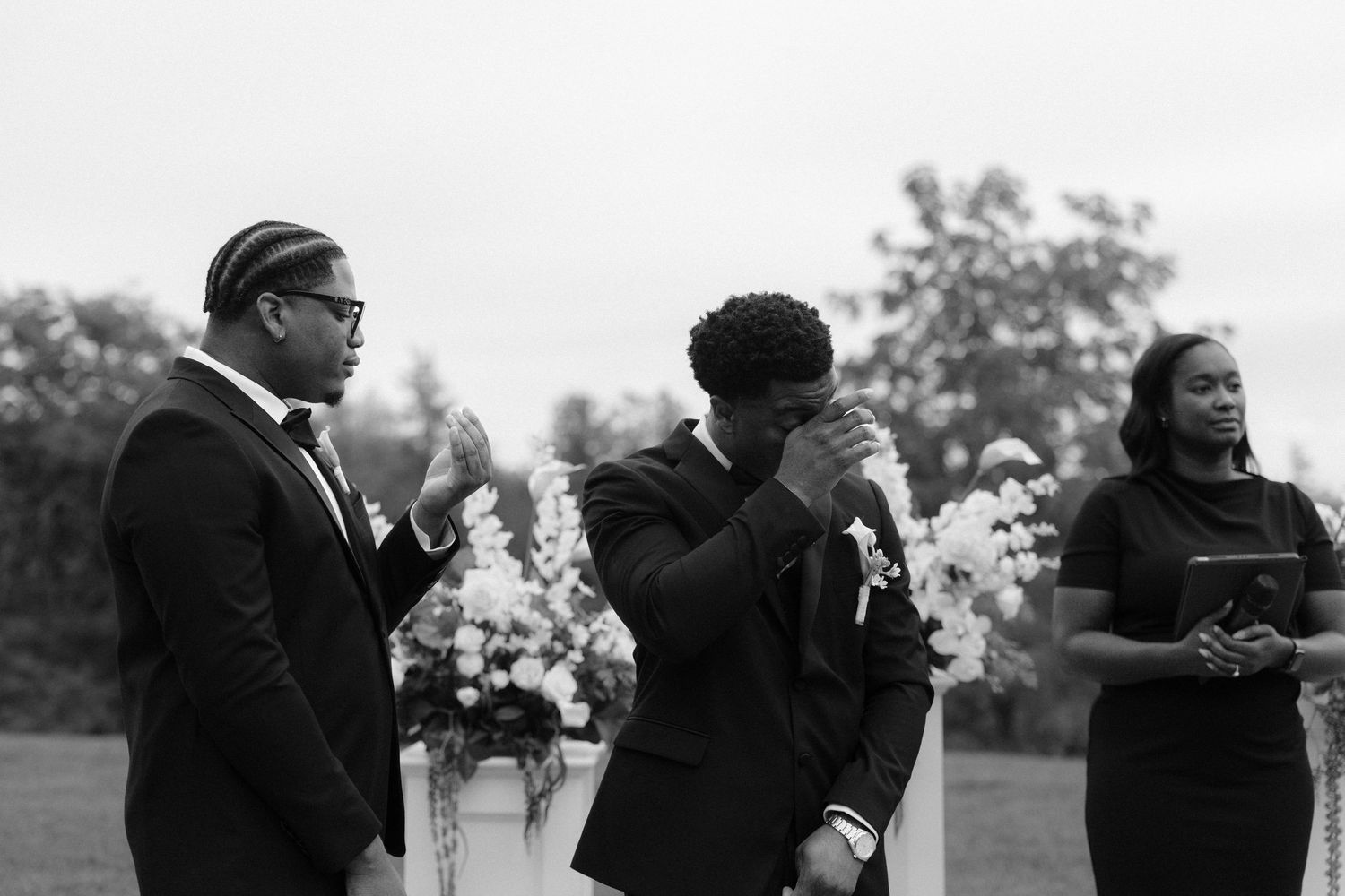 Emotional groom crying at outdoor wedding ceremony with officiant and bridesmaid standing nearby amid floral arrangements.