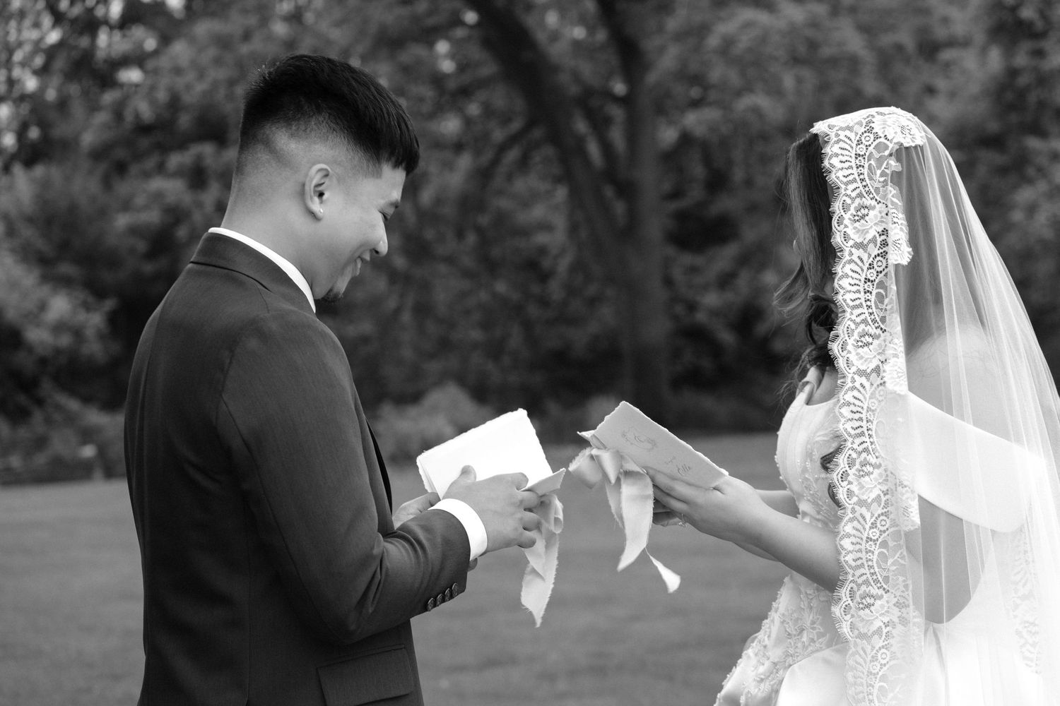 Black and white photo of a groom reading wedding vows to his veiled bride outdoors during a romantic ceremony.