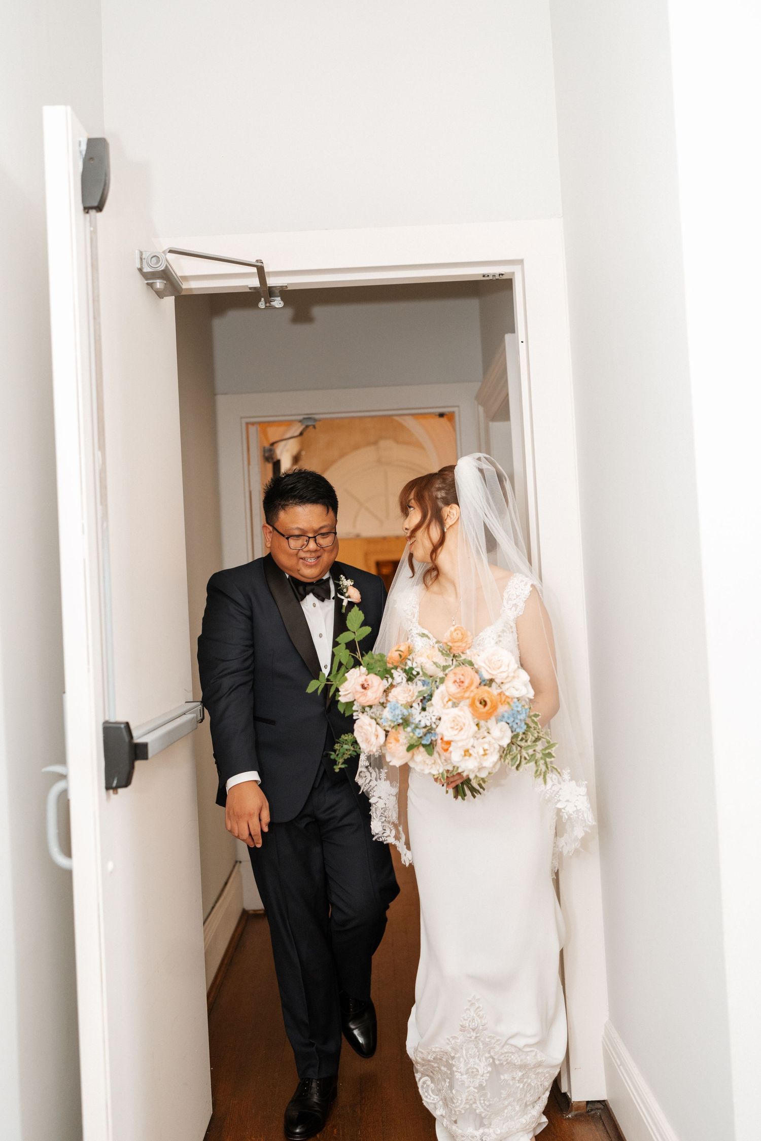 A couple walks through a narrow hallway wearing traditional wedding attire including a white gown and black tuxedo.