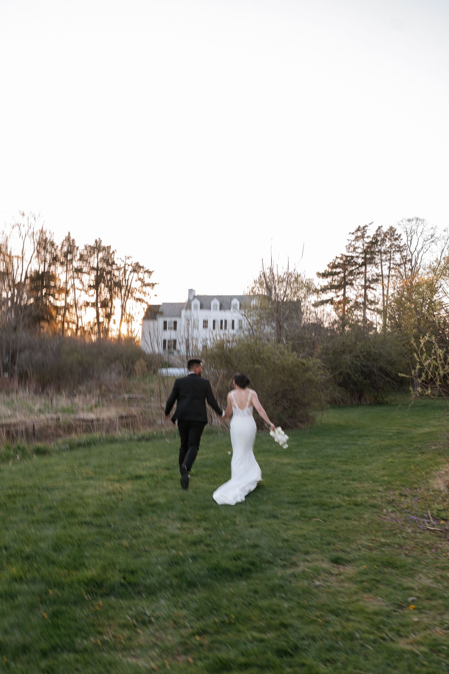 A couple walks hand in hand across a grassy field towards a white house at sunset with tall pine trees in the background.