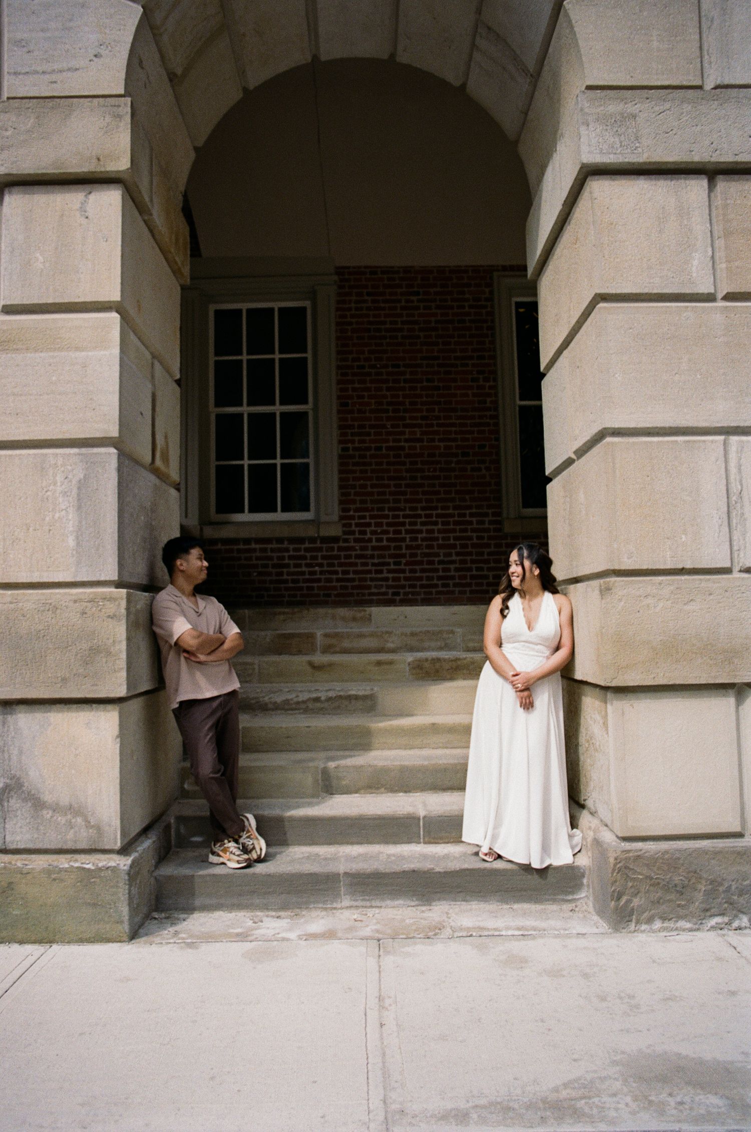 Couple poses on stone steps under arched doorway of historic brick building, bride in white halter dress.