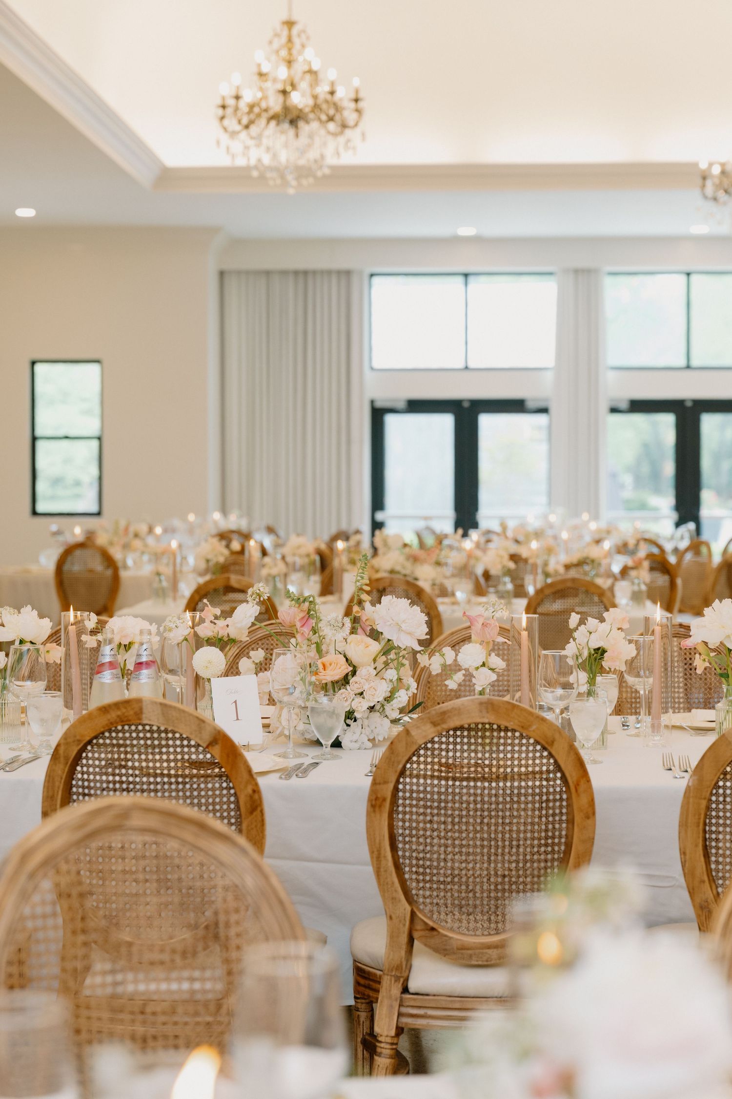 Elegant wedding reception hall with rattan chairs, white floral centerpieces, chandelier, and soft neutral tones.