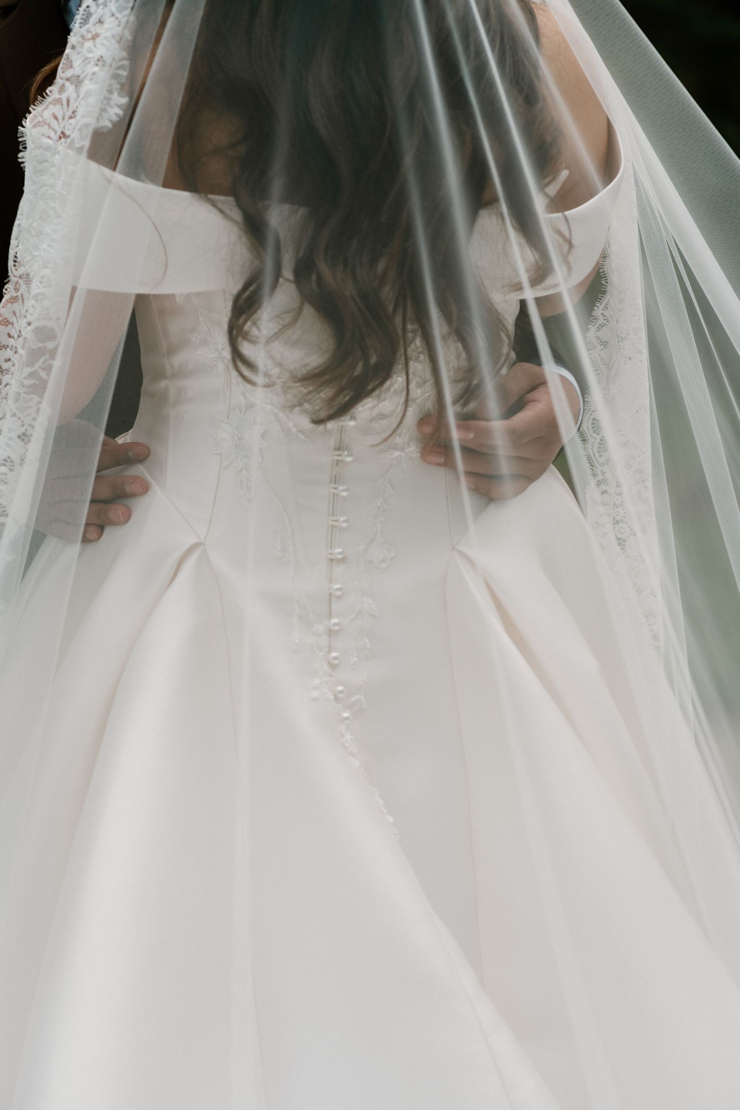 Bride in elegant white wedding gown with veil and button-back detail, viewed from behind.