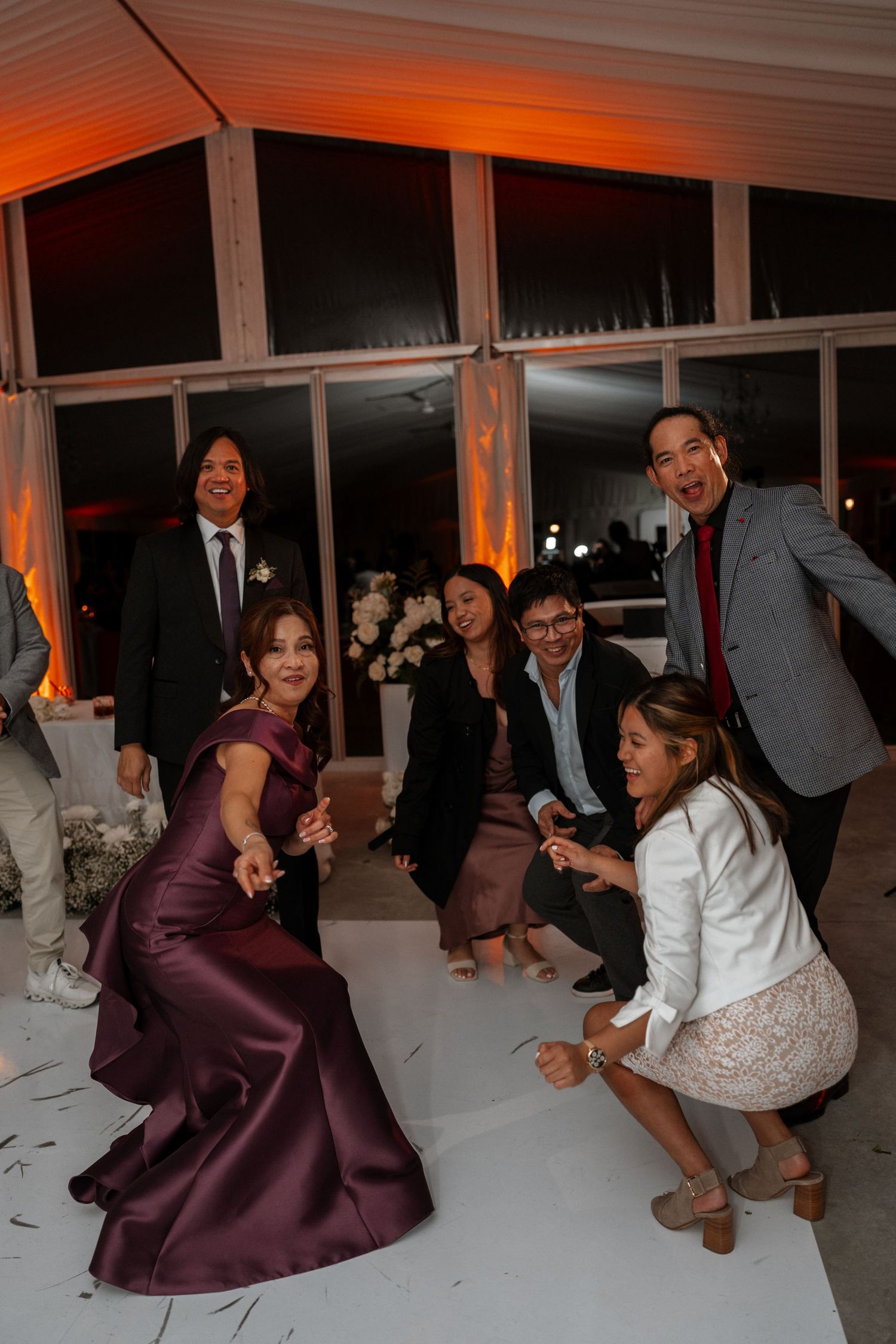 Guests in formal attire gather at an evening event with orange uplighting and elegant decor in a windowed venue space.