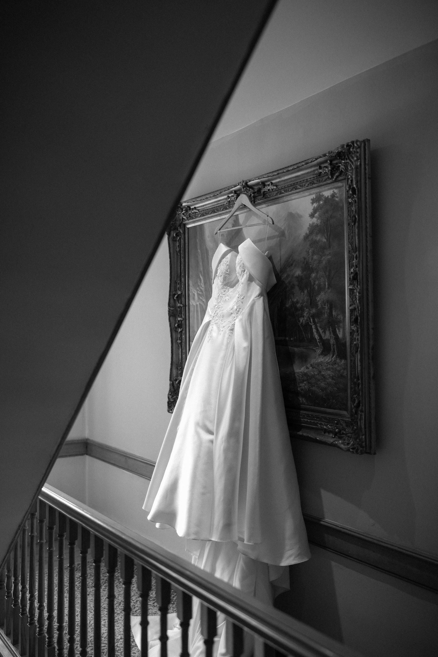 Black and white photo of an elegant wedding dress hanging on a ornate framed painting on a staircase wall.