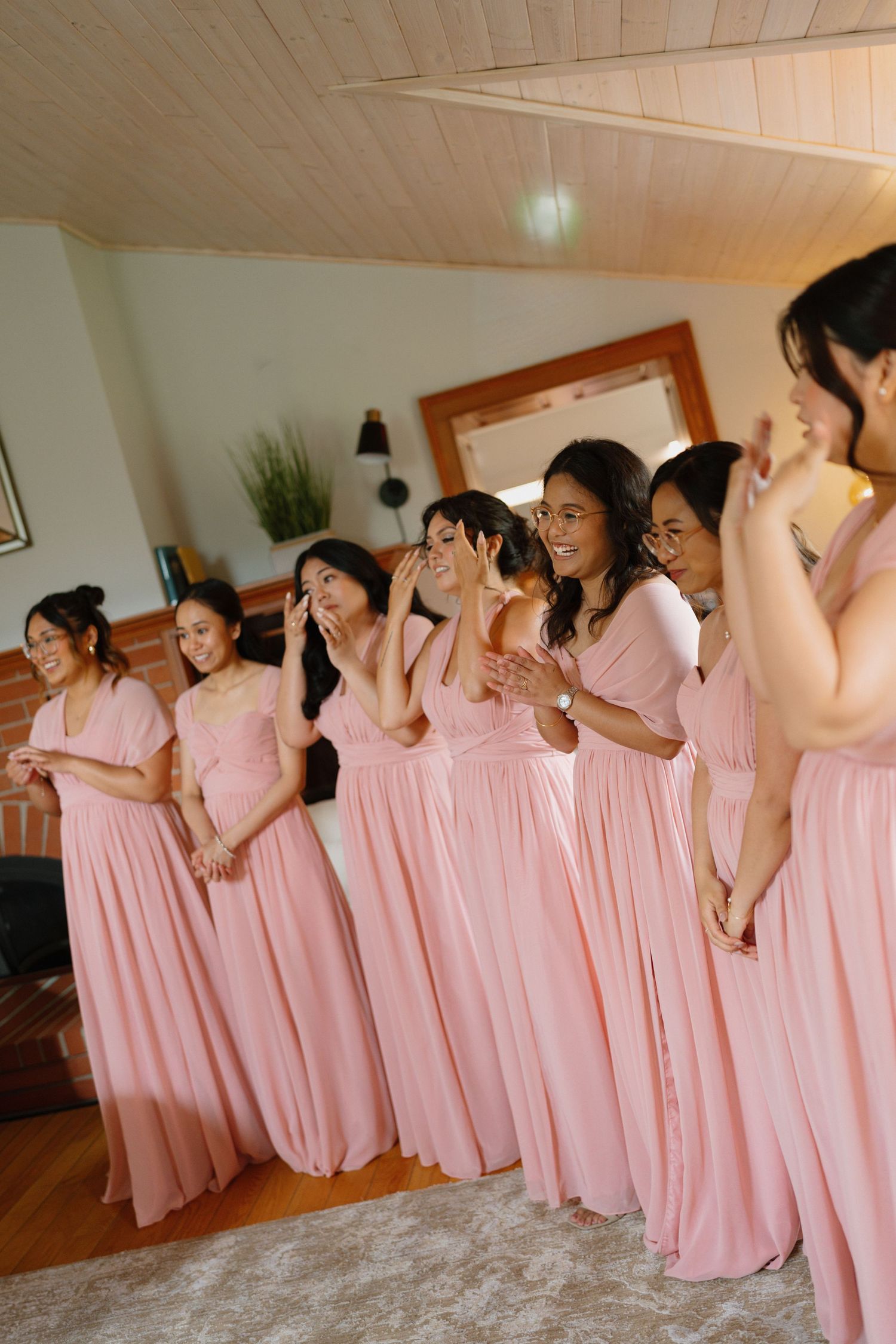 Bridesmaids in matching pink floor-length dresses laughing and reacting emotionally during wedding preparations indoors.