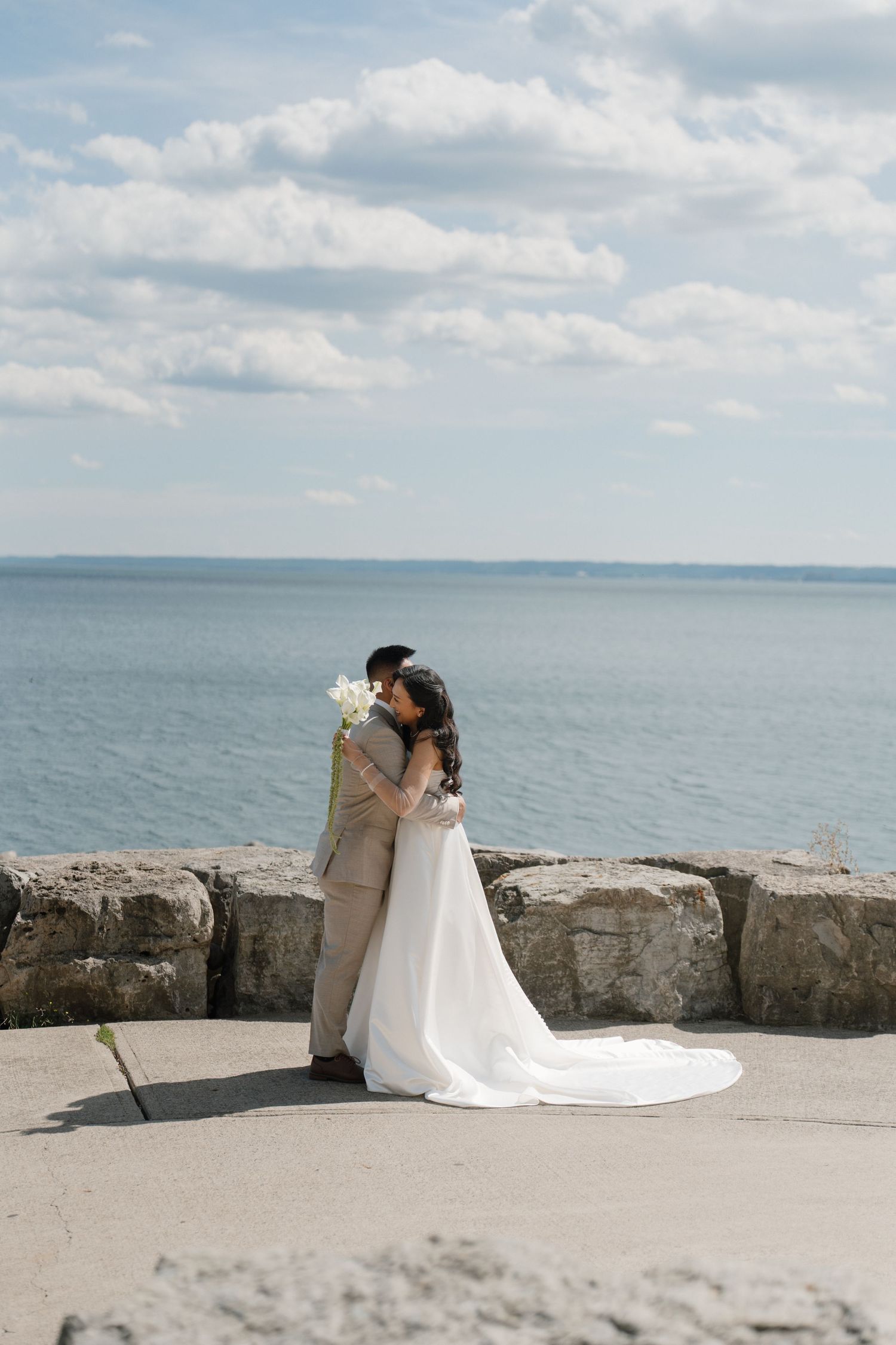 Bride and groom embrace near stone wall overlooking calm blue ocean under partly cloudy sky on their wedding day.