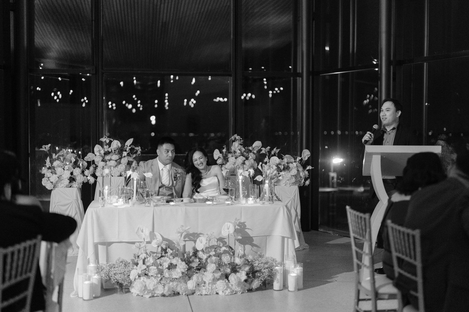 Black and white wedding reception photo showing couple at sweetheart table with floral arrangements and guest giving a toast.