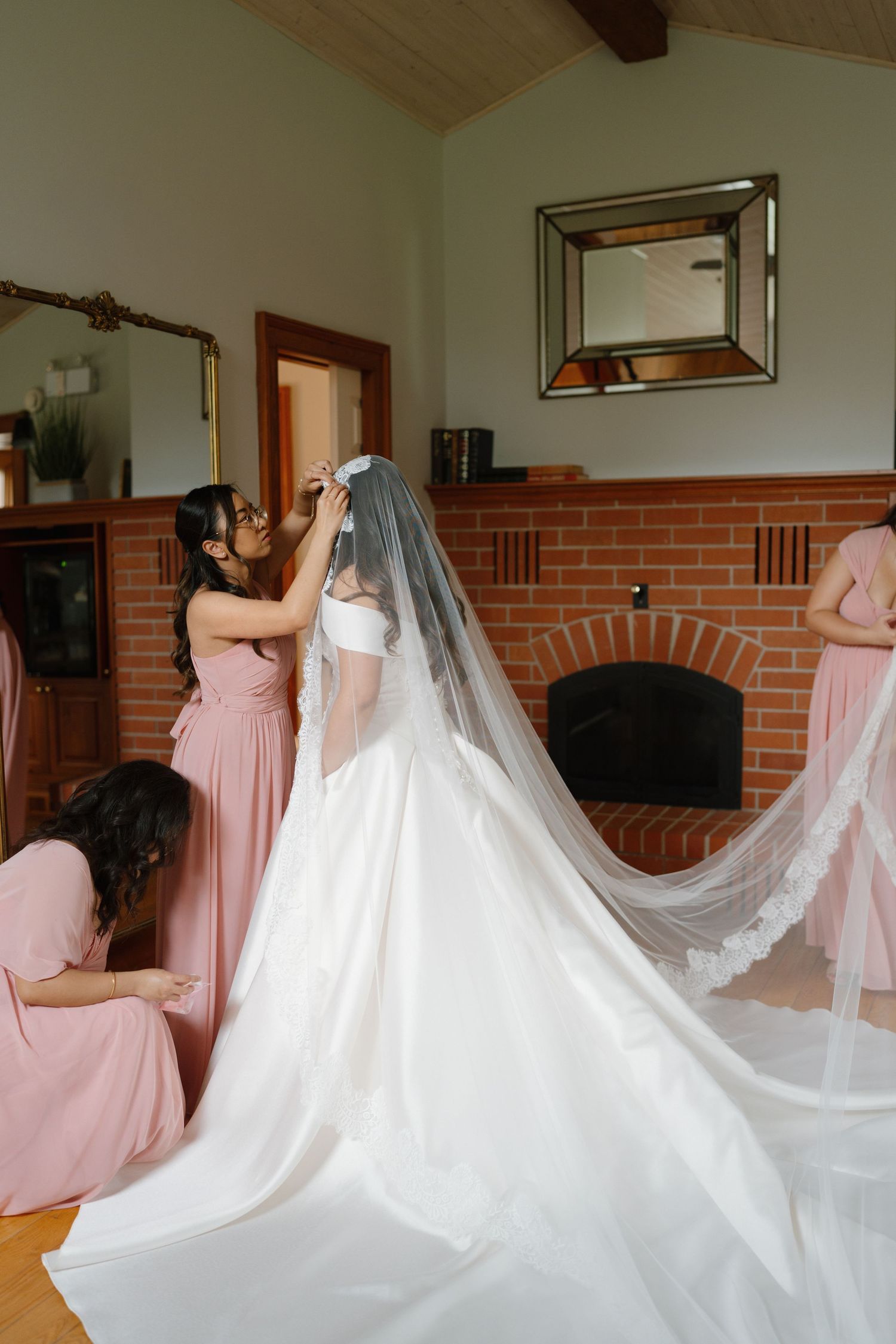 Bridesmaids in pink dresses helping bride with veil and train in a cozy room with brick fireplace.