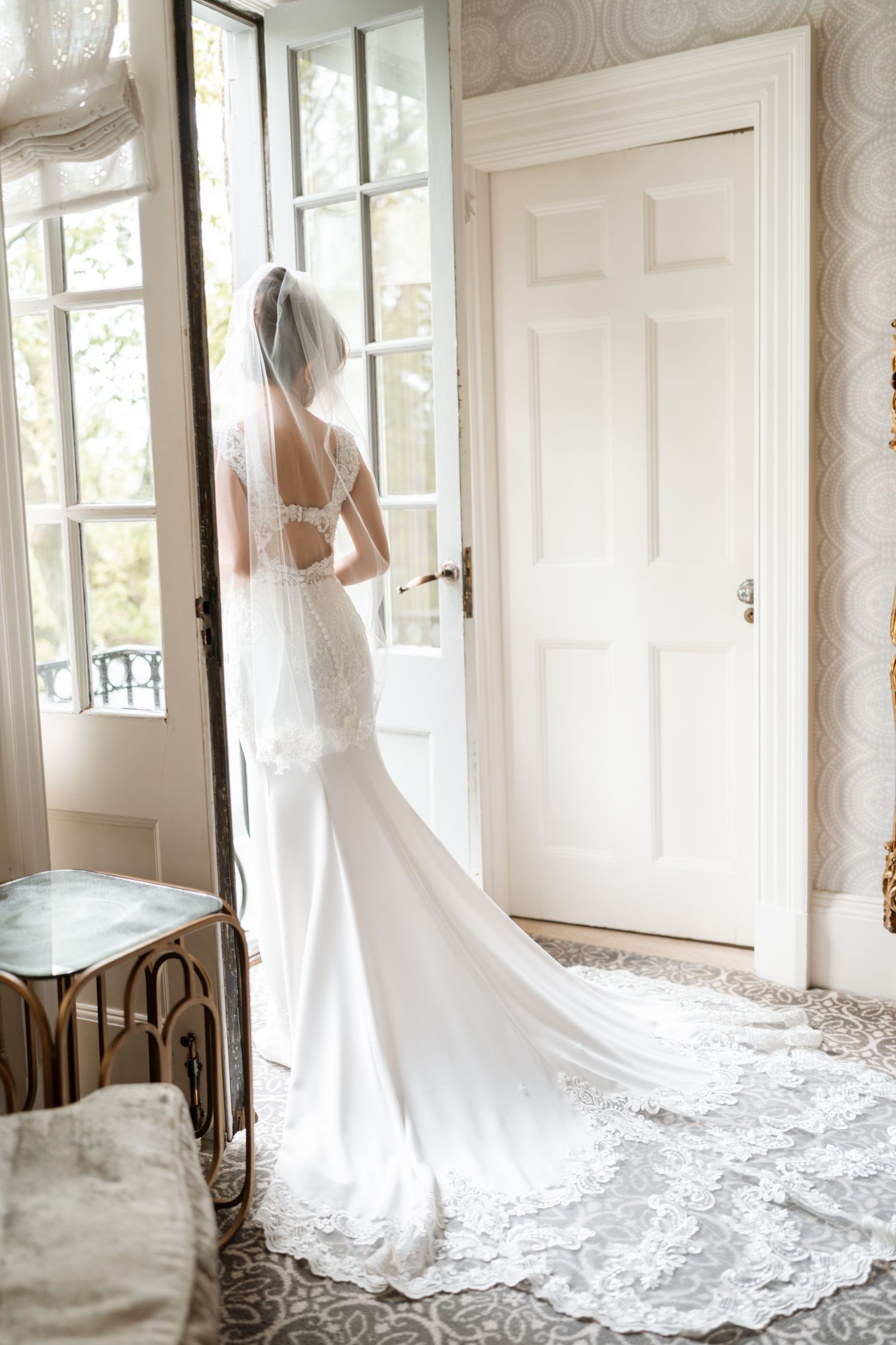 Elegant lace wedding dress with long cathedral train shown against white door and window in sunlit room.