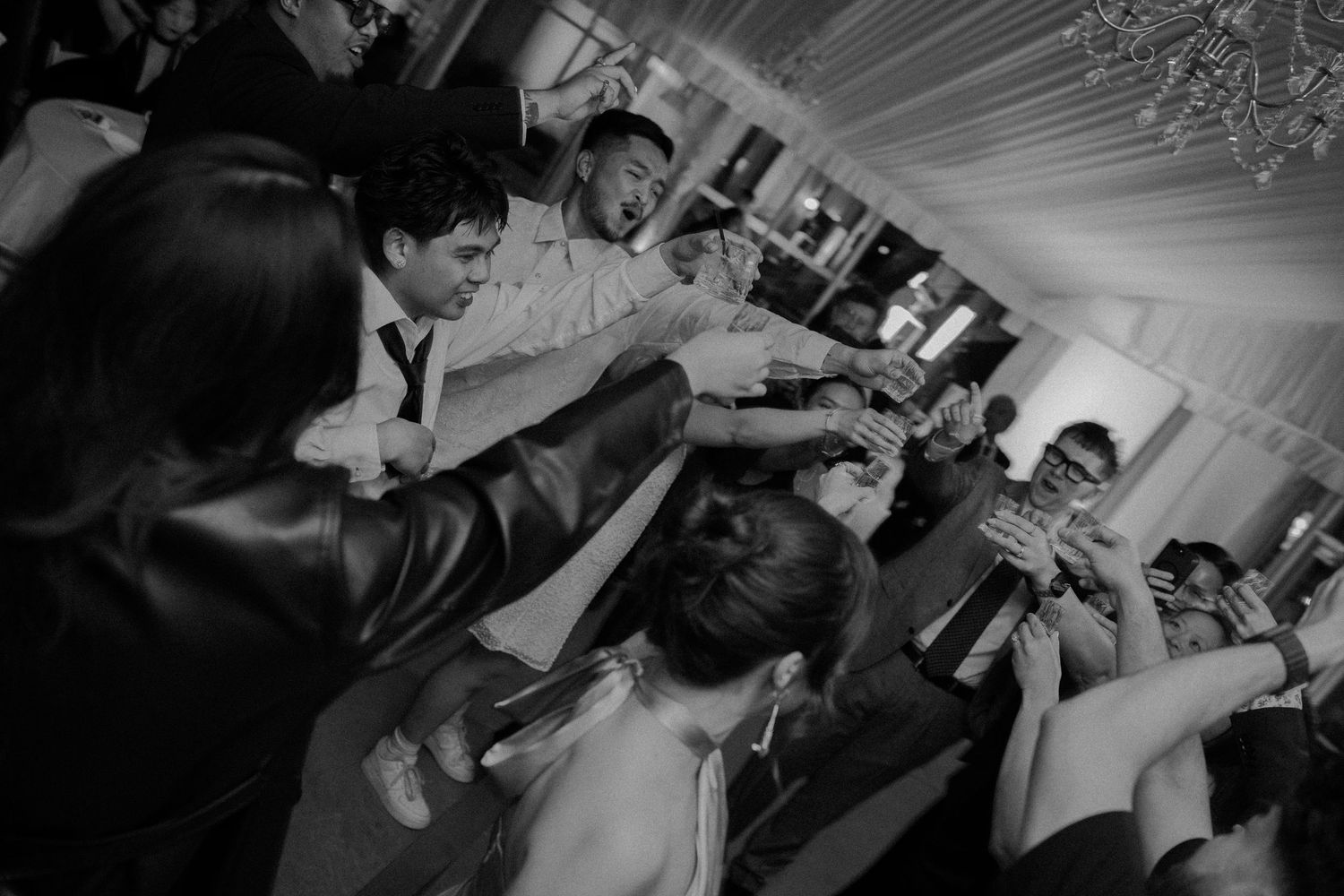 Party-goers raise their drinks in celebration at a wedding reception, creating an energetic black and white scene.